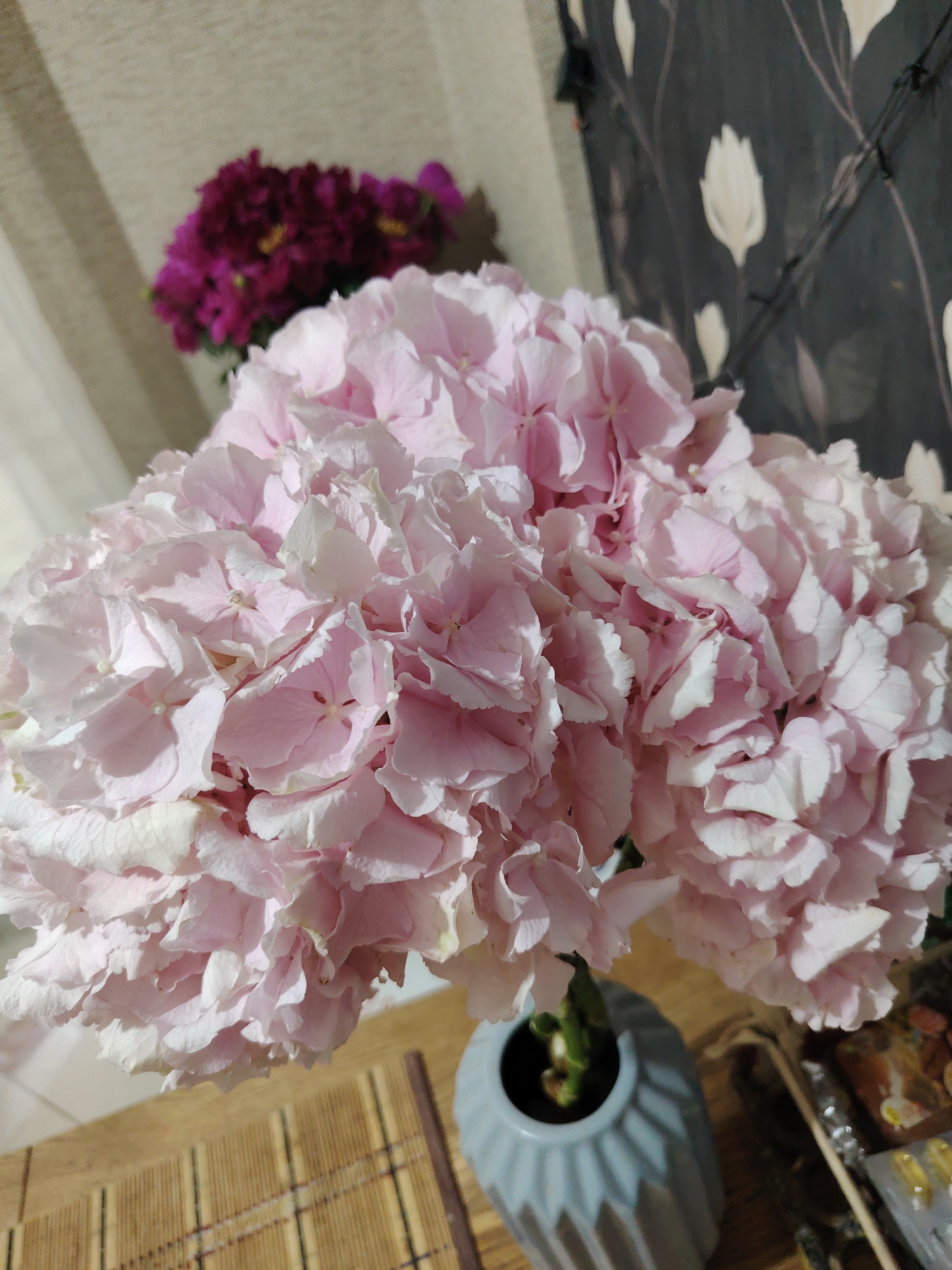 In a close-up, vertical shot, a large bouquet of light pink hydrangeas fills the foreground. The flowers are densely packed, with individual petals exhibiting delicate folds and soft lighting that creates subtle shadows. Behind the hydrangeas, a smaller, out-of-focus cluster of darker purple flowers is visible. The background features a textured, beige curtain on the left and a dark curtain with a subtle floral pattern on the right. The hydrangeas are placed in a pale blue, textured vase. The vase sits on a light brown, bamboo-patterned placemat. To the right of the vase, some indistinct items are present on a surface, including what appears to be a clear container with yellow capsules. The scene is indoors, with soft lighting suggesting it may be evening or a dimly lit room. The specific location within the city of Unknown, Unknown, is not determinable from the image.