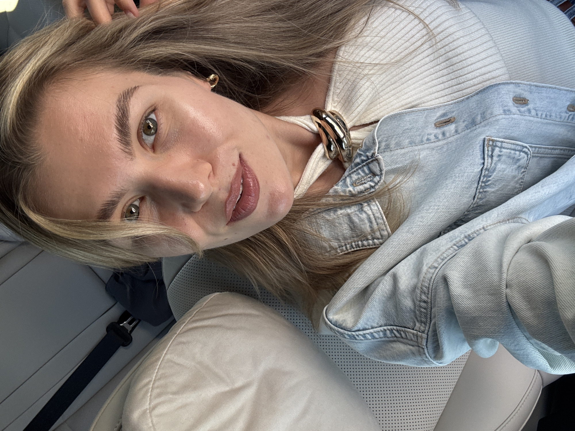 A young woman is taking a selfie from the backseat of a car. She has blonde hair and is wearing a denim jacket over a white ribbed top with a distinctive gold necklace. Her makeup is natural, with a focus on her lips. The interior of the car is visible, with light-colored leather seats and a seatbelt. The lighting suggests it is daytime.