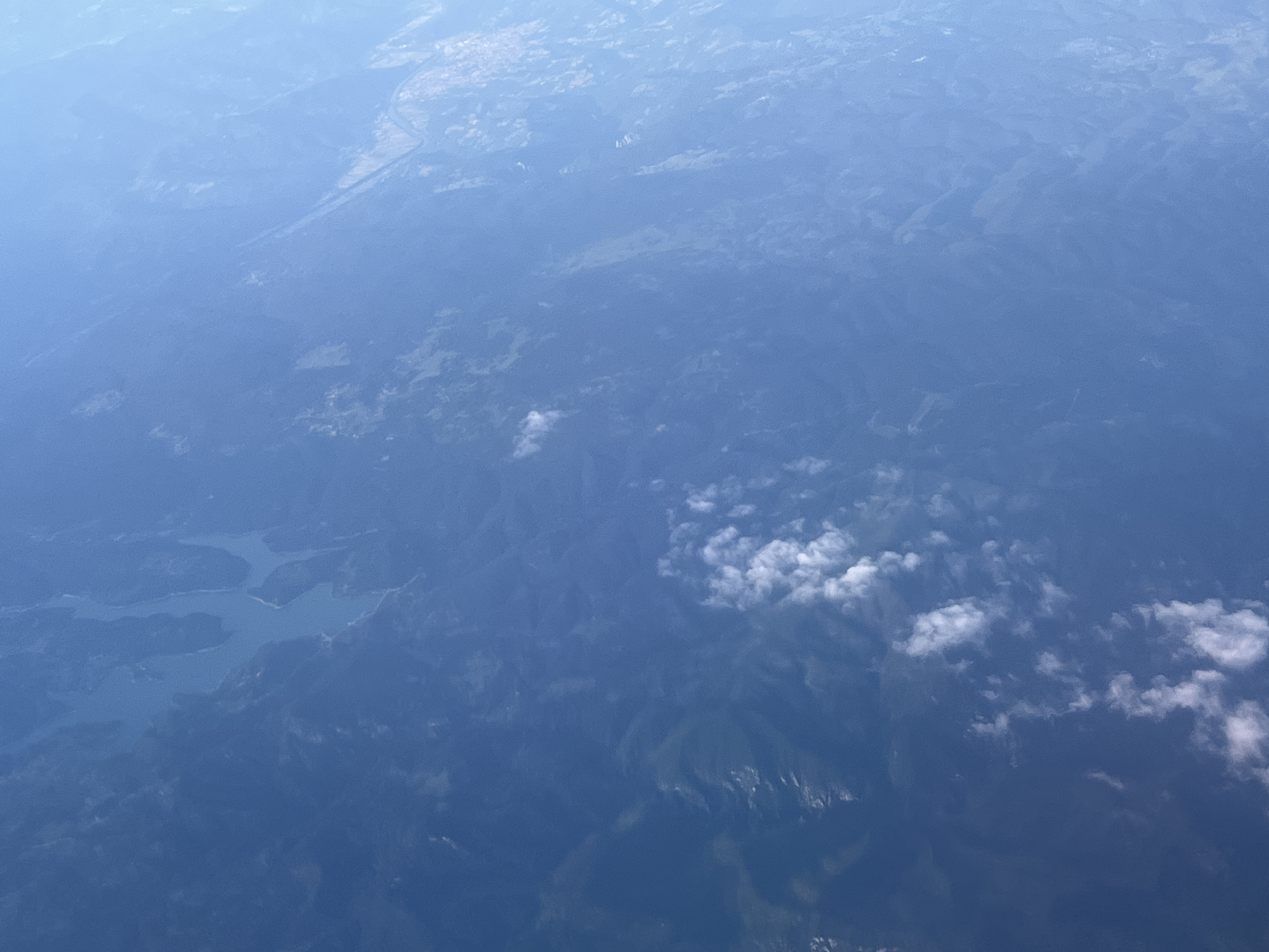 This is an aerial panoramic view, likely captured from an aircraft, showcasing a diverse landscape under a hazy blue sky.
The top-left quadrant prominently features an urbanized area, identified as the city of Sofia, Bulgaria. This region appears as a lighter, mottled texture with indistinct patterns suggesting buildings and infrastructure, transitioning from the top edge towards the center. A distinct linear grey feature, consistent with a major road or highway, traverses the upper-middle section of the image, leading into the urban expanse.
In the lower-left quadrant, a large, irregularly shaped body of water, definitively identified as the Iskar Reservoir, dominates the scene. Its light blue surface sharply contrasts with the surrounding land, revealing multiple arms and inlets.
The central, right, and lower portions of the image are dominated by extensive, undulating terrain characterized by dense, dark green vegetation, indicative of forested mountains or hills. Within these forested areas, scattered lighter green patches suggest clearings or areas with different vegetation types. Towards the bottom-right and central-right, higher elevations with brighter, patchy white areas are visible, potentially representing snow or exposed rock on mountain peaks.
Atmospheric conditions include a pervasive light blue haze across the entire vista. Several scattered, distinct white cumulus clouds are present, primarily in the middle and lower-right sections, casting subtle shadows upon the terrain below. No individual people, specific vehicles, or direct human interactions are discernible given the high altitude.