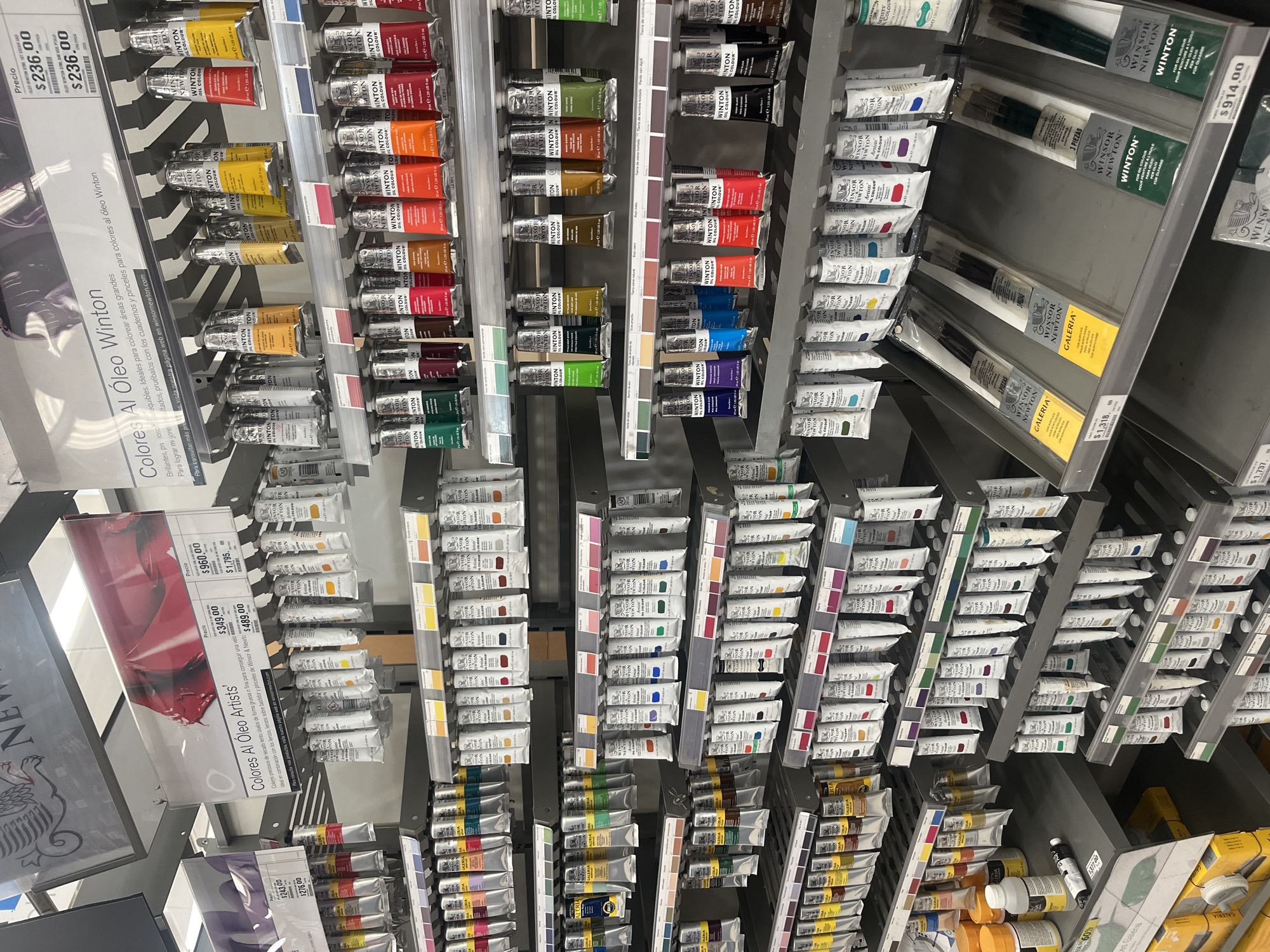 The image shows shelves stocked with oil paints and brushes at an art supply store, likely in Puebla, Mexico. The shelves are arranged vertically, displaying a variety of brands and colors of oil paints, including Winsor & Newton Winton and Artists' lines. Prices are displayed on shelf tags in Mexican pesos, such as $236.00, $349.00, $960.00, and $1,795.00. The store also carries sets of Winsor & Newton brushes. The image focuses on the product displays, providing a detailed view of the inventory and pricing. The environment is a typical retail setting for art supplies. There are no people in the frame.