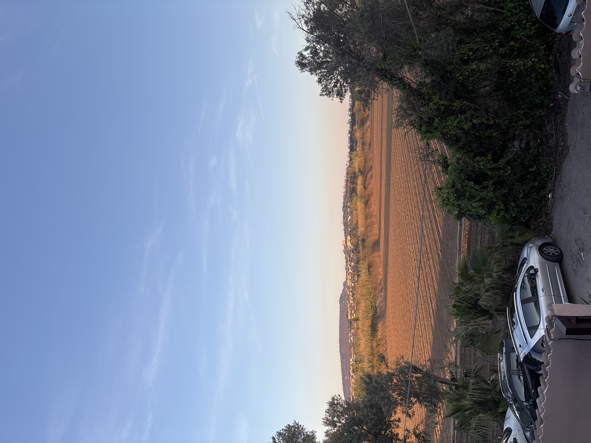 The image presents a landscape view taken from an elevated position, possibly a balcony. The right side of the image features a partially visible silver car parked near vegetation, including palm trees. A paved area suggests a road or driveway. To the left, the scene opens up to a view of agricultural land with distinct rows, indicating tilled soil. A line of buildings is visible in the distance, suggesting a populated area. The sky above transitions from a pale blue to a light orange hue, indicative of either sunrise or sunset. The presence of trees along the edge of the frame further frames the scene. The setting appears to be Giugliano in Campania, Italy, based on the landscape and architectural characteristics.