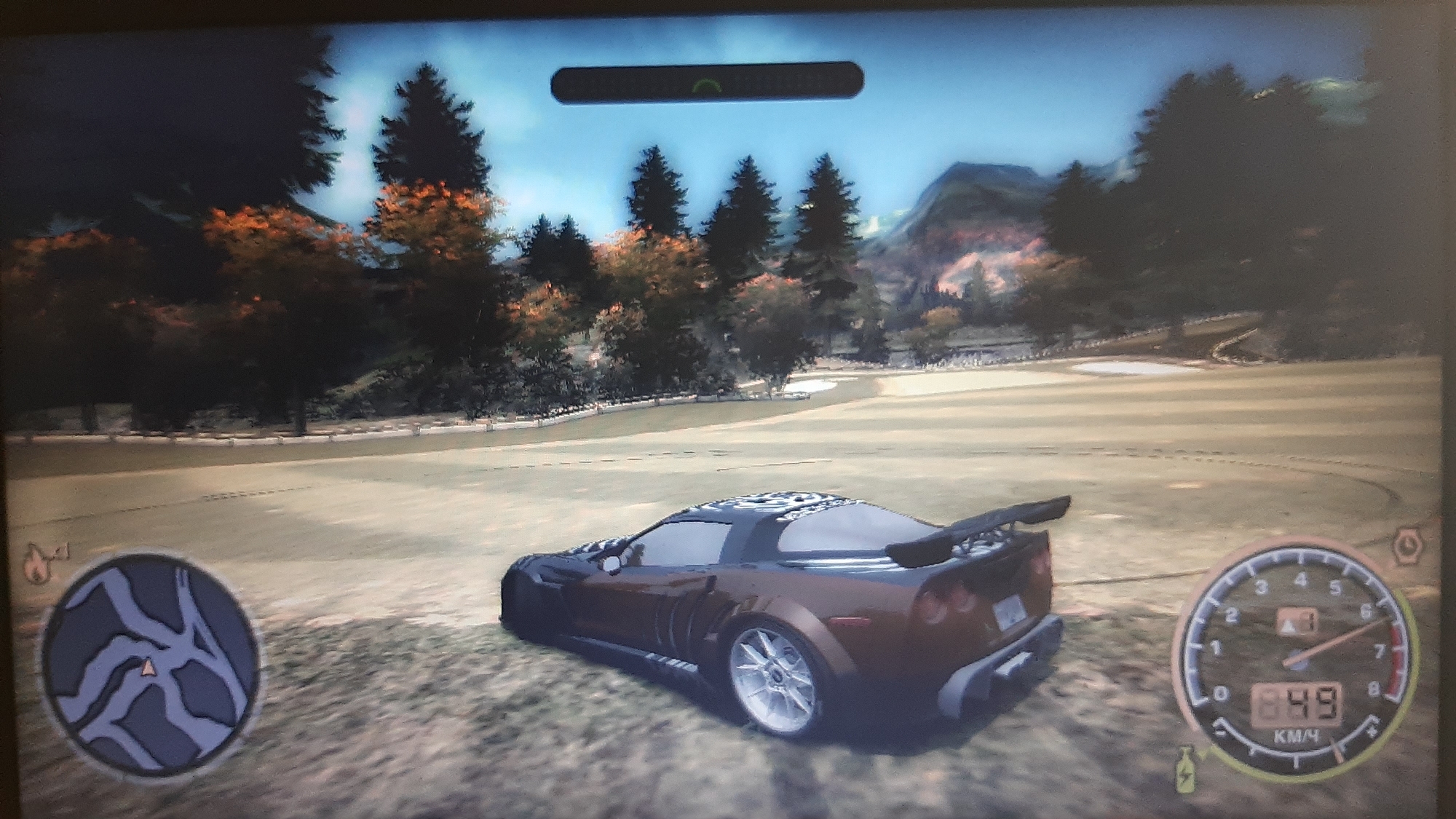 The image shows a screenshot from a racing video game. A dark brown sports car with a spoiler is parked on a grassy area, possibly a golf course, in a mountainous, wooded environment. The car has a custom decal on its roof. In the lower left corner, a circular mini-map shows a road network and a marker. In the lower right corner, a speedometer displays a speed of 89 km/h and a fuel gauge. A horizontal bar with a green indicator is visible at the top of the screen, suggesting a game progress or power-up indicator. The background features trees with autumn foliage and distant mountains under a clear blue sky.