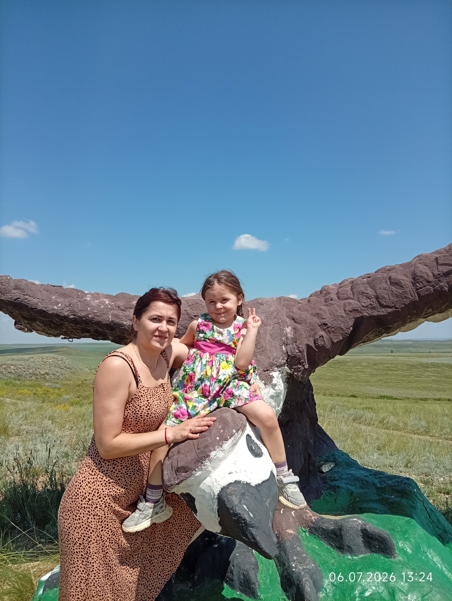 A woman and a young girl are posing for a photo outdoors. The woman is wearing a brown leopard-print dress and is seated on a large, brown, bird-like sculpture. The girl, wearing a colorful floral dress, is sitting on the sculpture's back, with her legs dangling. She is holding up two fingers in a peace sign. The sculpture appears to be situated in a grassy, open area with rolling hills in the background under a bright blue sky with a few clouds. The location is identified as Aleksandrovka, Kazakhstan, and the timestamp on the image reads 06.07.2026 13:24.