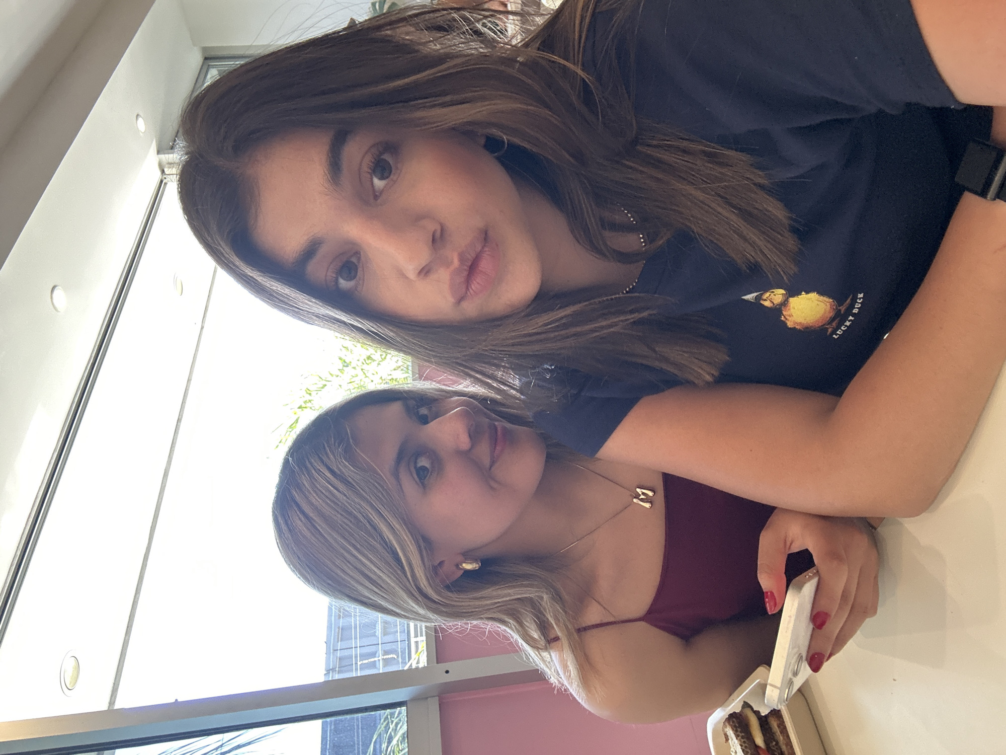 Two young women are depicted indoors, likely within a cafe or restaurant in Puebla, Mexico. The woman in the immediate foreground, positioned to the right, has long dark brown hair and wears a navy t-shirt with a "LUCKY DUCK" graphic. She has a black digital watch on her left wrist and looks directly at the camera with a slight pout. To her left, slightly behind and in the mid-ground, a woman with long blonde hair wears a burgundy spaghetti strap top, gold hoop earrings, and a necklace with a prominent "M" pendant. She is seen smiling subtly towards the camera, looking over her right shoulder. Her right hand, featuring red painted fingernails, holds a white smartphone. A white table surface extends across the foreground. On the table to the left, a white rectangular container holds a layered food item. The background reveals large windows offering natural light, with glimpses of outdoor foliage and indistinct building structures. An interior pink wall panel is visible behind the woman with blonde hair. The ceiling features recessed light fixtures.