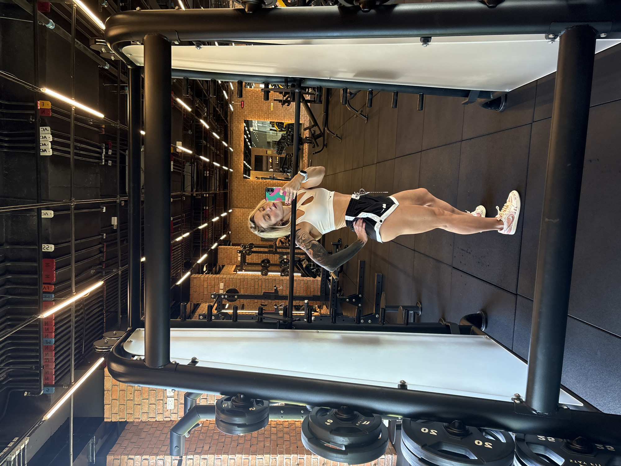 In a gym located in Rawai, Thailand, a woman with blonde hair and tattoos on her arms is taking a selfie in a large mirror. She is wearing a white cutout sports bra and black shorts with white trim. The gym features exposed brick walls, industrial-style lighting, and various weightlifting equipment, including a squat rack and weight plates. The flooring is black rubber. The woman is standing next to a squat rack with weight plates stacked on the side. The reflection in the mirror shows the gym's interior with other workout machines and a brick wall with a large mirror.