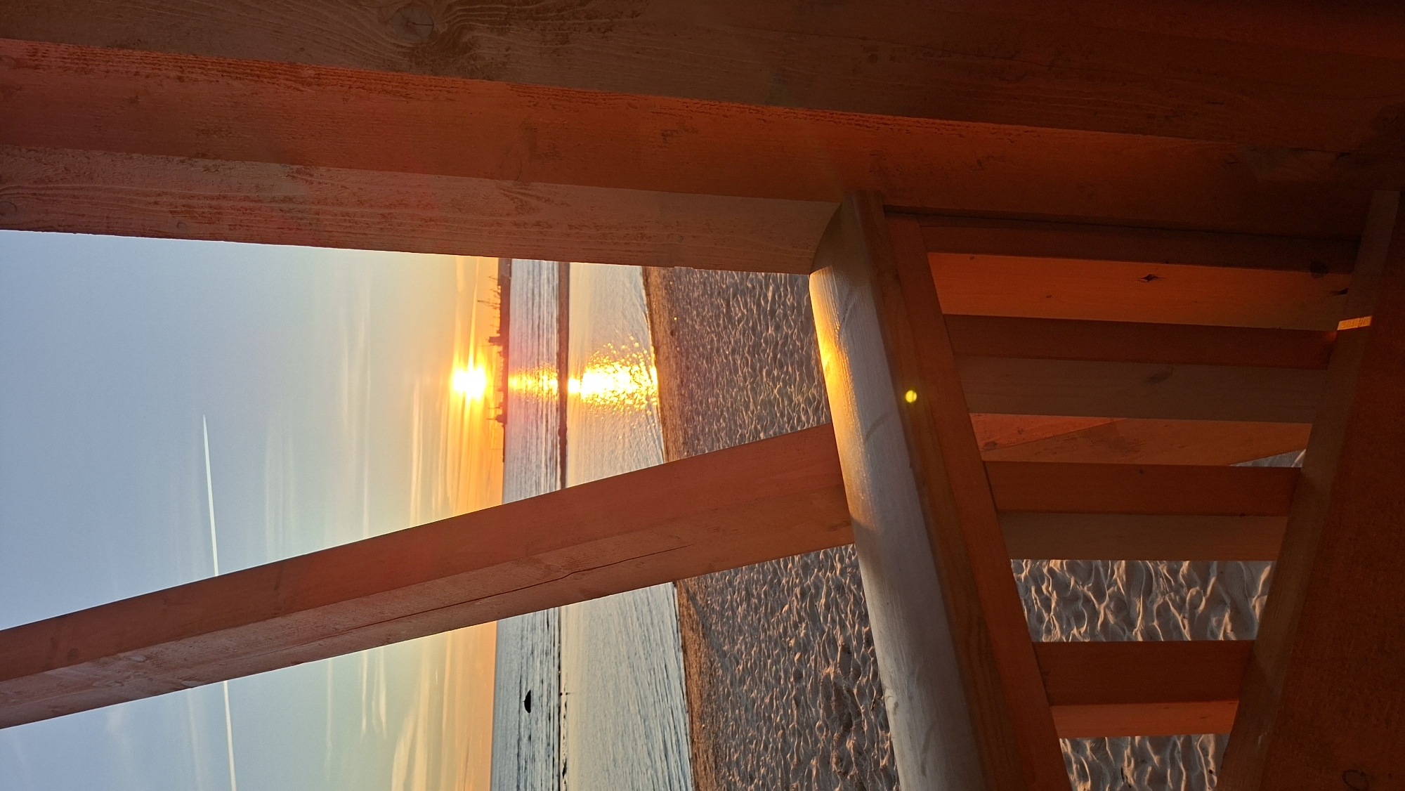 The image depicts a sunset over the Baltic Sea, viewed from a wooden structure on a sandy beach. The sun, a bright orange orb, is low on the horizon, casting a shimmering reflection on the water. Silhouettes of ships and industrial structures are visible in the distance. A contrail from an airplane cuts across the clear blue sky. The foreground is dominated by the warm-toned wooden beams and railings of a beach structure, illuminated by the setting sun. The sand on the beach appears fine-grained and is marked by gentle ripples. The scene suggests a peaceful, serene moment at a coastal location. Given the presence of industrial silhouettes and the angle of the sun, this could be the coast near the Port of Tallinn, Estonia, where industrial activity and the sea meet.