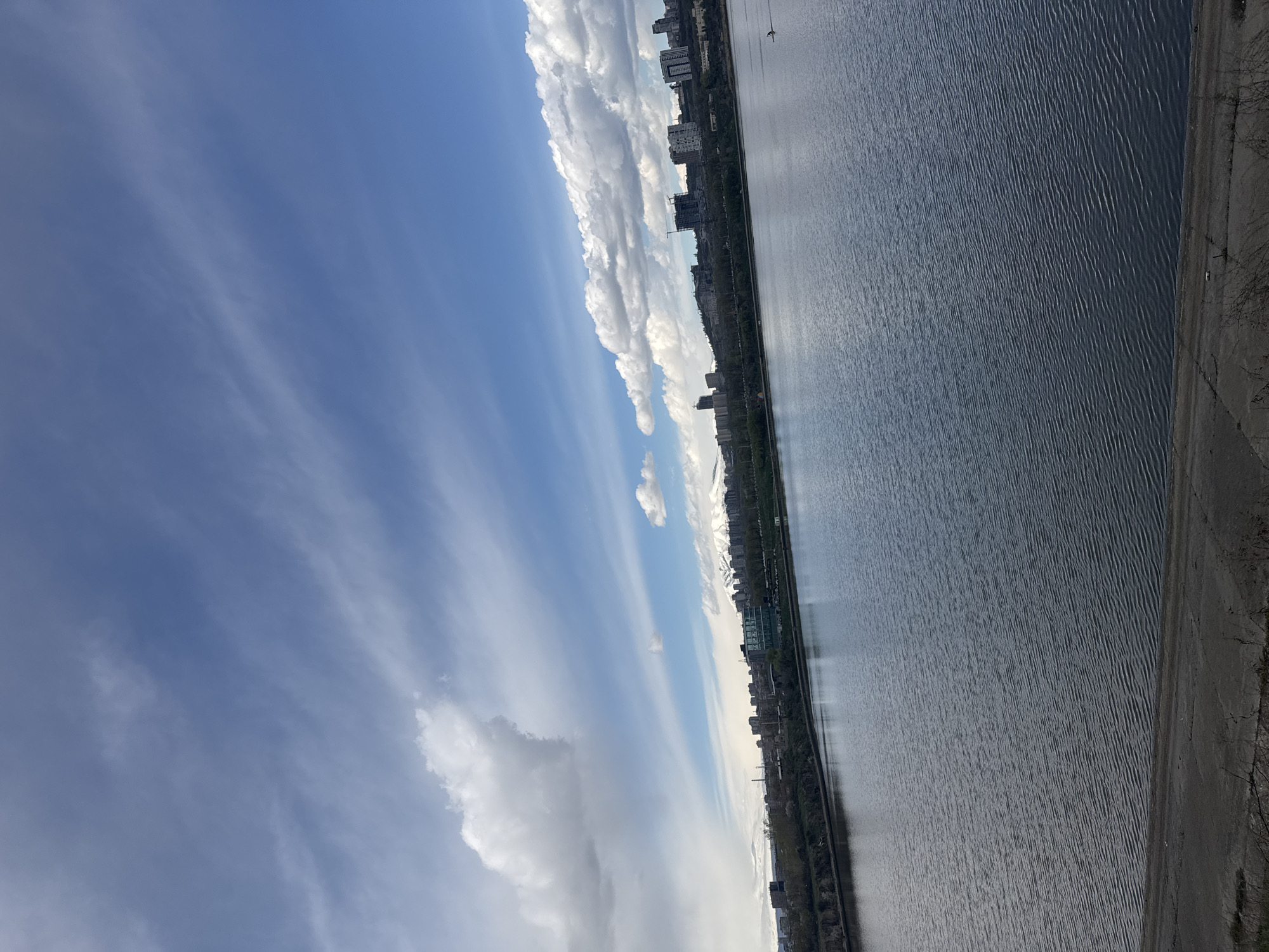 The image depicts a cityscape alongside a large body of water, likely a river or lake, under a blue sky with scattered clouds. In the distance, buildings of varying heights can be seen lining the waterfront. The water surface exhibits a rippled texture, suggesting a slight breeze. The photograph is taken at an oblique angle, giving a perspective of the city as it stretches along the water's edge. The location is not Yerevan, Armenia.