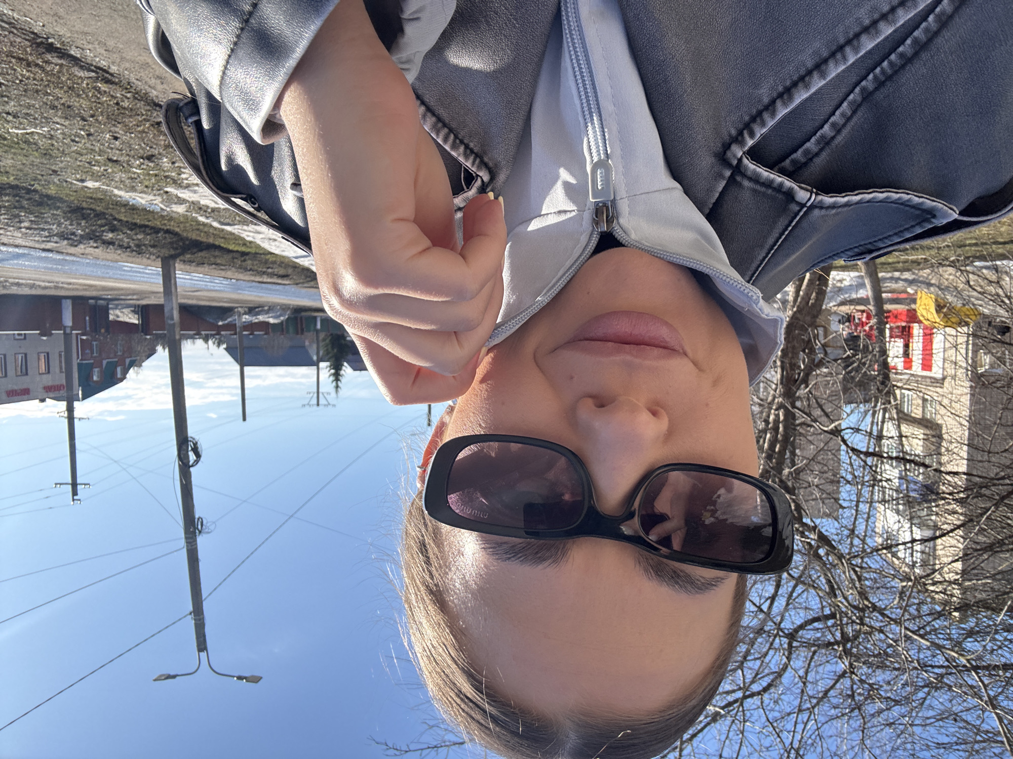 A selfie of a fair-skinned woman wearing dark sunglasses and a gray jacket is oriented upside down. The background reveals a cityscape, including buildings, a playground with red and yellow elements, and bare trees. Utility poles and wires stretch across a clear blue sky. The image appears to have been taken outdoors in Chernushka, Russia based on available information.