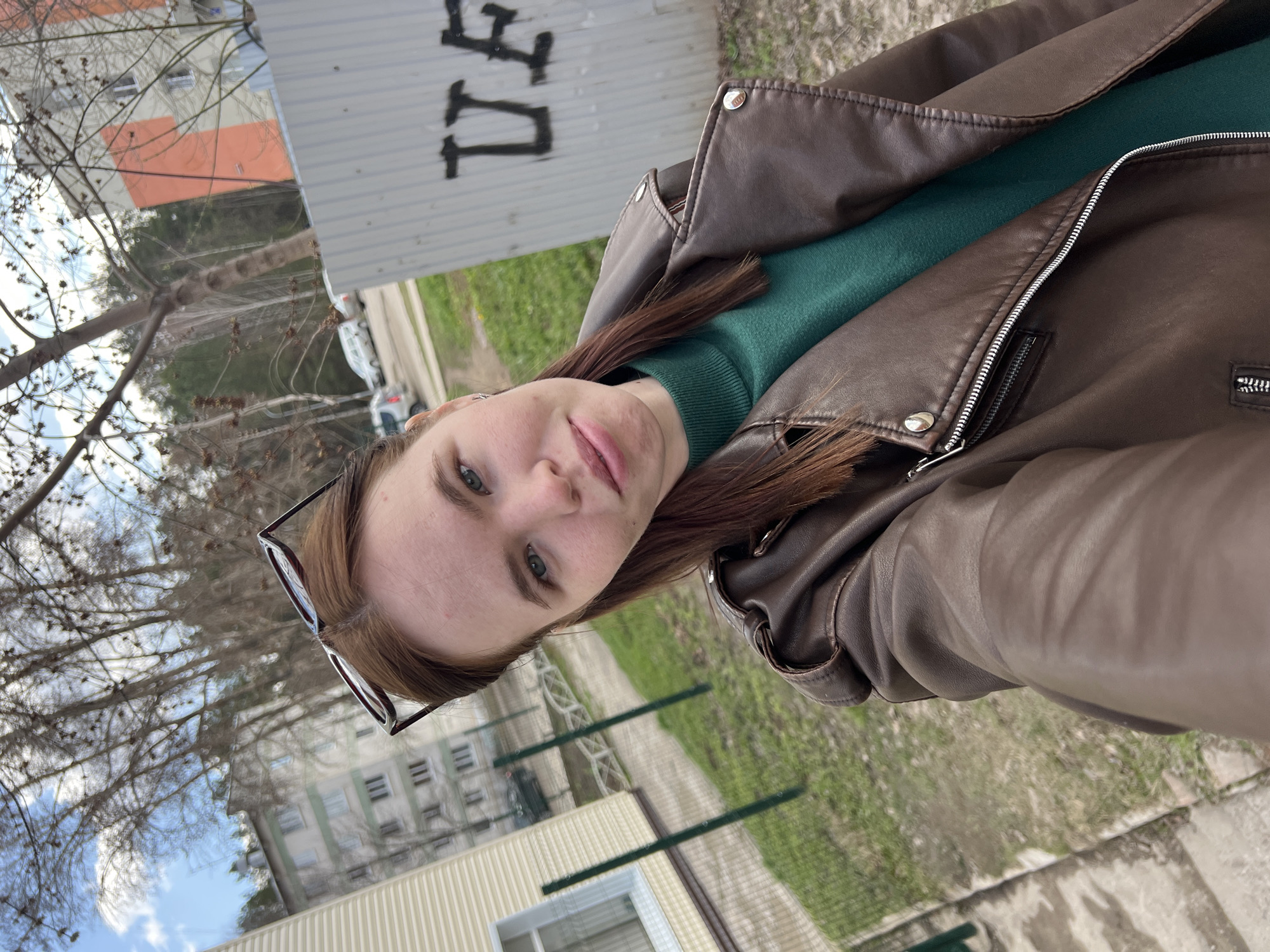 A young woman, appearing to be in her late teens or early twenties, is depicted in a selfie-style portrait. She has fair skin, light blue-green eyes, and long, reddish-brown hair. Her expression is neutral as she looks directly at the camera. She is wearing a dark brown faux leather jacket with silver zippers and snap buttons, layered over a dark green turtleneck sweater. Black-framed sunglasses are perched on top of her head.
The setting appears to be outdoors in an urban or suburban environment, likely in Chaikovsky, Russia. The background features several bare, leafless trees, indicating either late winter or early spring. Beyond the trees, there are multi-story residential buildings. To her right, a light-colored corrugated metal fence or wall is visible, with dark graffiti consisting of Cyrillic letters, possibly "ДЕЛ". To her left, a light-colored building with horizontal siding and a window can be seen. The ground consists of a mixture of grass, dirt, and asphalt, with a green metal fence separating areas. In the distance, several cars are parked. The sky is partly cloudy, with patches of blue visible, and the lighting suggests it is daytime, possibly late morning or early afternoon.
