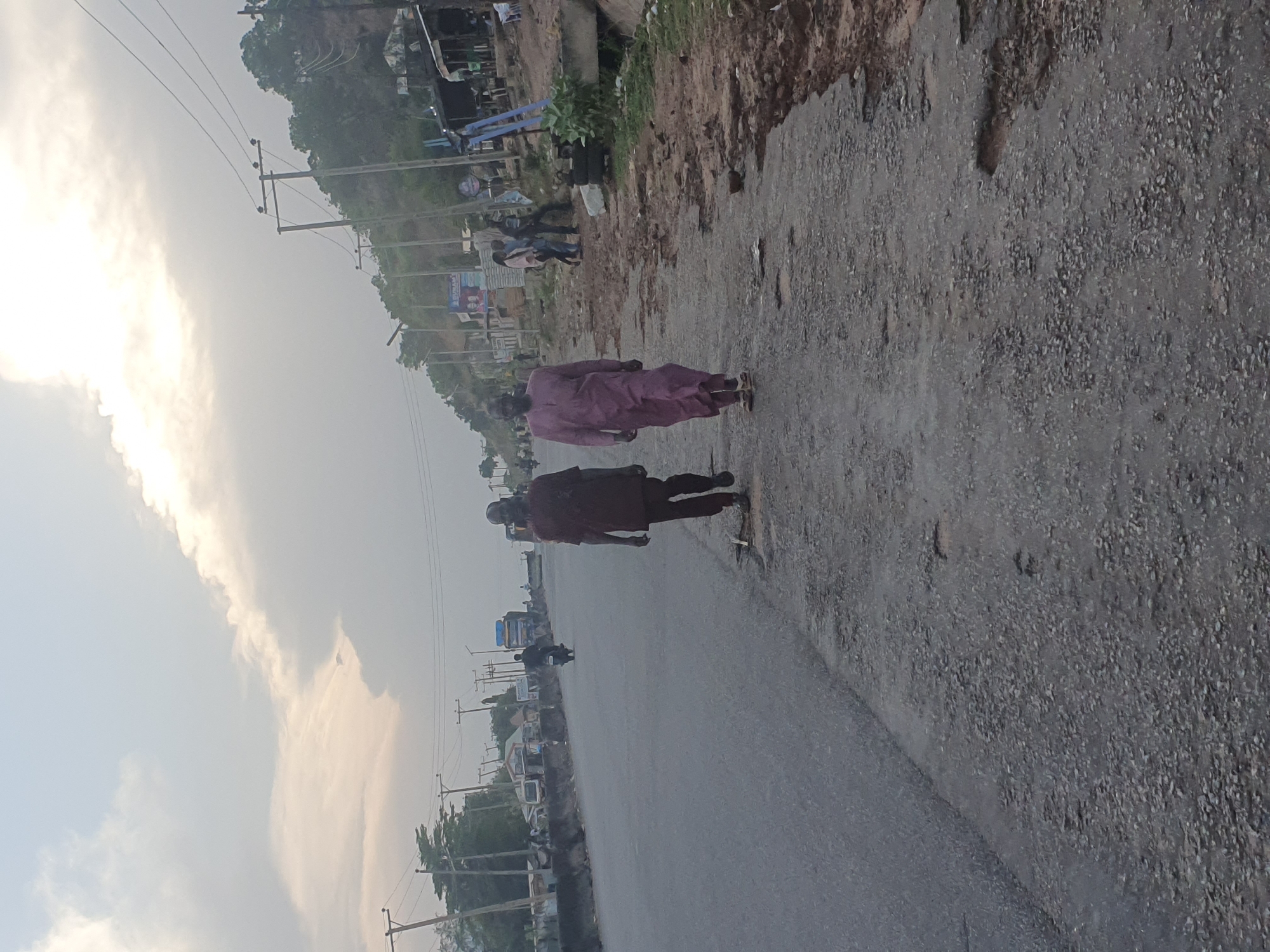 The image depicts a road in Lokoja, Nigeria. Two people walk along the shoulder of the road. One individual wears a mauve-colored robe, while the other wears a dark brown robe. A motorcycle is visible on the road, as are several vehicles further in the distance. Power lines run along the roadside. The environment includes a hillside with structures and vegetation. The sky is overcast.