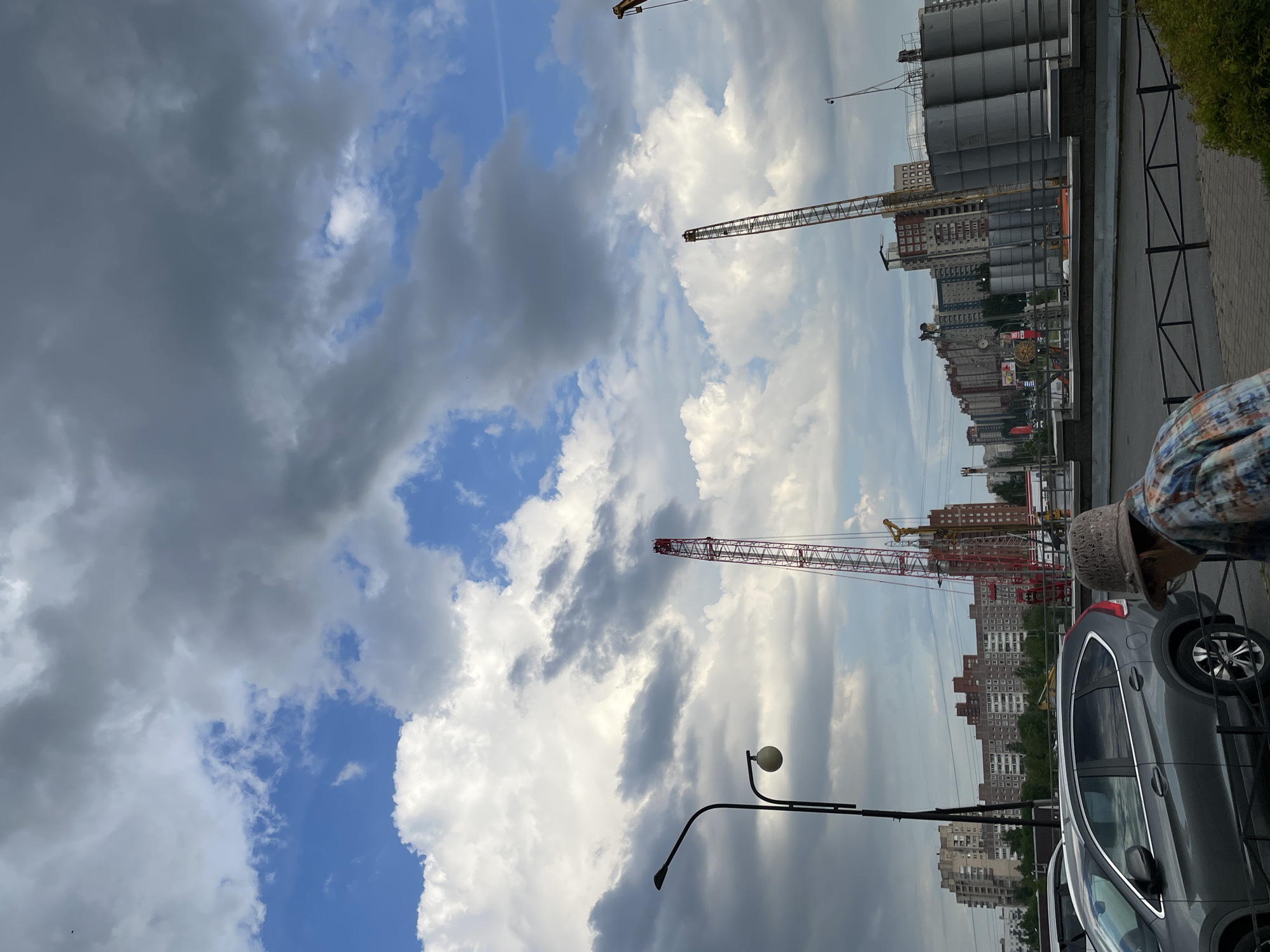 This image captures a vibrant urban construction scene under a dynamic sky, set in what appears to be Saint Petersburg, Russia. The sky occupies a significant portion of the frame, displaying a striking contrast between large, heavy grey clouds and areas of clear blue sky dotted with bright white cumulus clouds, suggesting a variable weather condition, possibly in the late afternoon or early evening.
In the mid-ground, the skyline is punctuated by at least two prominent tower cranes: one in yellow/orange and another in red, indicating ongoing building activity. Below these cranes, numerous multi-story buildings, many appearing to be residential or commercial, extend across the scene. Some structures are clearly under construction. Notably, a series of large, grey, cylindrical silos or storage tanks are visible on a raised platform, hinting at industrial facilities integrated within the urban sprawl.
The foreground features elements of a typical street environment. A black metal fence runs along the bottom edge, separating the viewer's perspective from the background. A modern, dark grey SUV is partially visible on the right, parked alongside the fence. A person, whose head and upper torso are visible on the far right, is wearing a light-colored hat and a multi-colored, patterned shirt. A black street lamp with two spherical lights extends into the lower-middle of the frame. Some green foliage is also visible behind the fence on the right. There is no visible text in the image. The overall scene depicts an active, developing cityscape.