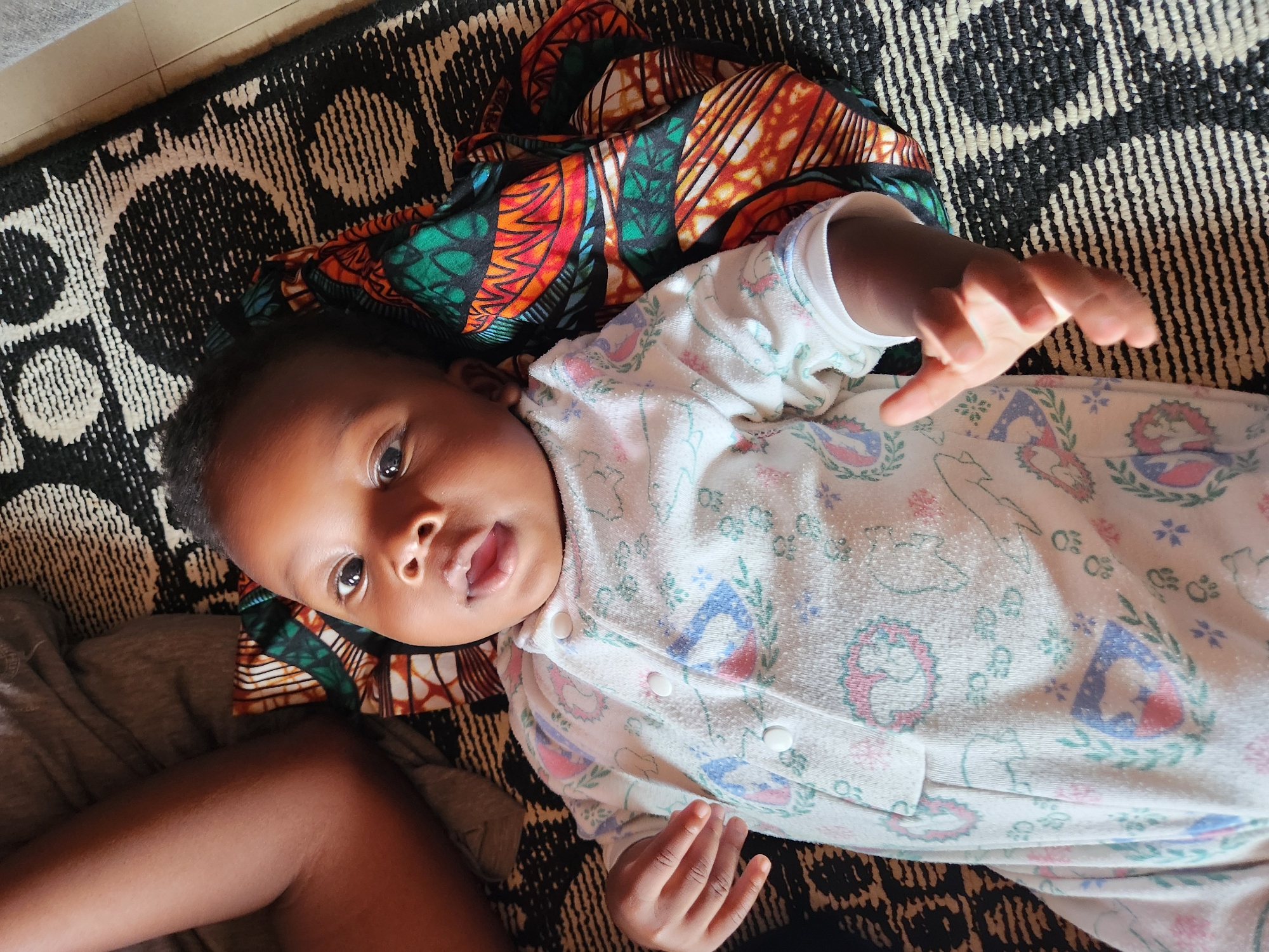 A high-angle, eye-level shot shows a baby lying on its back on a patterned rug. The baby is wearing a white onesie with pink and blue accents. To the baby's right, a brightly colored African print fabric is folded. The baby's left leg and foot are visible, as is a portion of an adult's leg to the upper left of the frame. The scene is indoors, in Sabon Tasha, Nigeria.