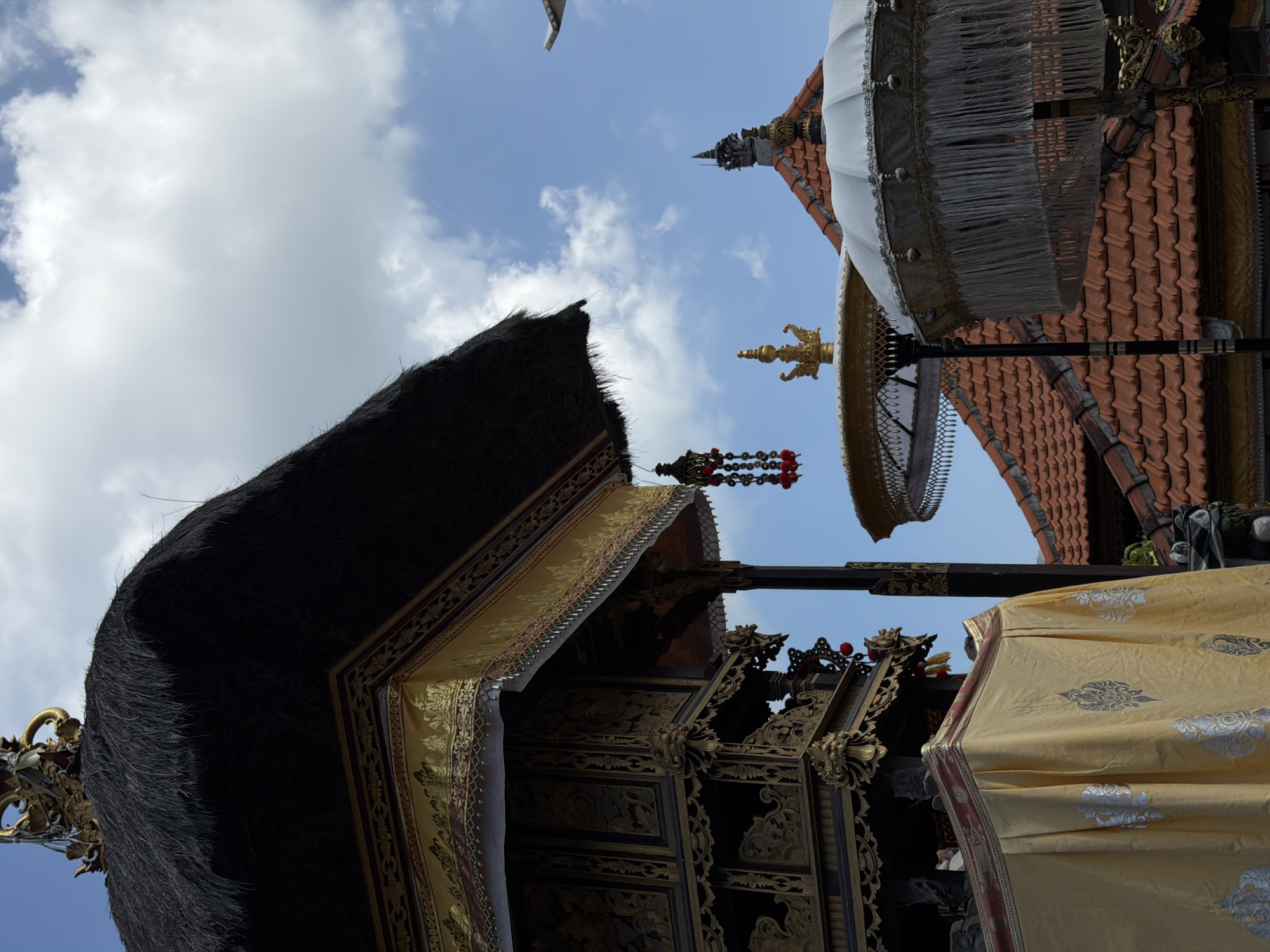 The image captures an outdoor scene in North Kuta, Indonesia, featuring ornate traditional Balinese architecture against a bright blue sky with scattered clouds. In the foreground, a thatched roof structure, adorned with intricate gold detailing and dark wooden carvings, dominates the left side of the frame. A yellow fabric draped over a portion of this structure displays a repeating silver pattern.
To the right of the main structure, several tiered roofs covered in reddish-orange tiles are visible. Two decorative umbrellas, one white with fringe and the other gold with intricate detailing, stand prominently. One of these umbrellas is partially obscured by the thatched roof. Hanging from the ornate structure is a decorative pendant with red elements. The overall impression is of a culturally rich religious or ceremonial site. No people are visible in the image.