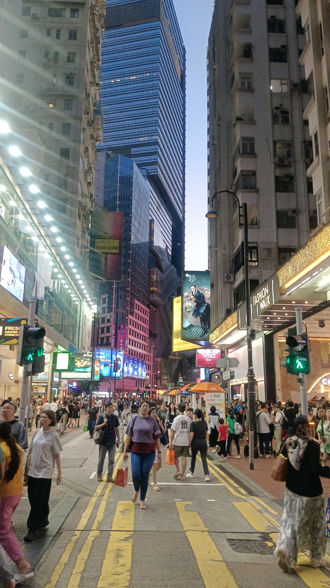 This image depicts a bustling street scene in Hong Kong, China, likely in a commercial district, possibly Causeway Bay or Mong Kok, given the density of high-rise buildings, neon signs, and pedestrian traffic. The perspective is from street level, looking down a wide avenue lined with towering buildings on both sides. The architecture is a mix of modern skyscrapers with glass facades and older, more utilitarian apartment buildings with visible air conditioning units and laundry lines.
The street is filled with a diverse crowd of pedestrians, suggesting a busy shopping area. People are walking in various directions, some alone, others in small groups. Their clothing styles indicate a range of ages and fashion preferences, with many carrying shopping bags. Several are looking at their phones, while others appear to be engaged in conversation. The presence of traffic lights, specifically a green pedestrian signal, indicates an active intersection.
Prominently featured are numerous large digital billboards and illuminated signs advertising various products and services. These signs contribute to the vibrant and dynamic atmosphere of the city. A particularly noticeable sign displays a man in a suit holding food items. Another sign has the text "FASHION WALK" with illuminated golden accents, suggesting a shopping destination. Orange umbrellas are visible, possibly indicating street vendors or temporary stalls.
The road surface is asphalt, marked with thick yellow double lines, typical of pedestrian crossings. The lighting suggests it is either early evening or late afternoon, with a clear sky visible between the buildings. The overall impression is one of urban density, commercial activity, and a lively street life characteristic of Hong Kong.