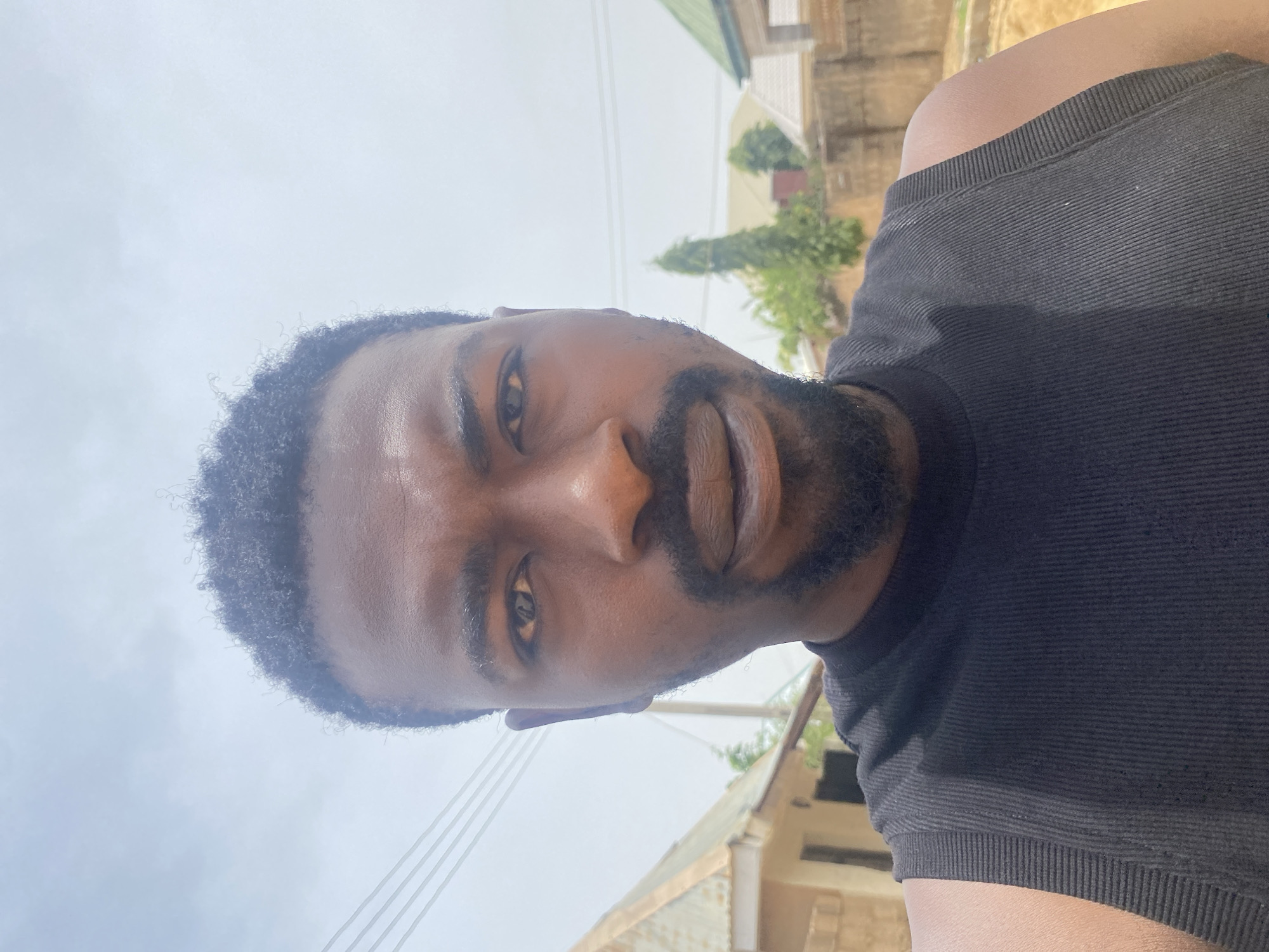 A close-up selfie, oriented on its side, depicts a man with dark skin, dark curly hair, and a full beard and mustache. He looks directly at the camera with a neutral expression. He is wearing a dark, likely black or dark gray, ribbed sleeveless top.
The image is captured outdoors during bright daylight, under a hazy or overcast sky, providing soft, diffuse lighting. The background, visible on the right side of the frame, includes parts of light-colored buildings with some green roofing, along with green foliage from trees or bushes. Thin utility lines are also visible against the bright sky. The setting appears to be a residential or semi-urban area, consistent with Barakin Akawo, Nigeria. No text is visible in the image.