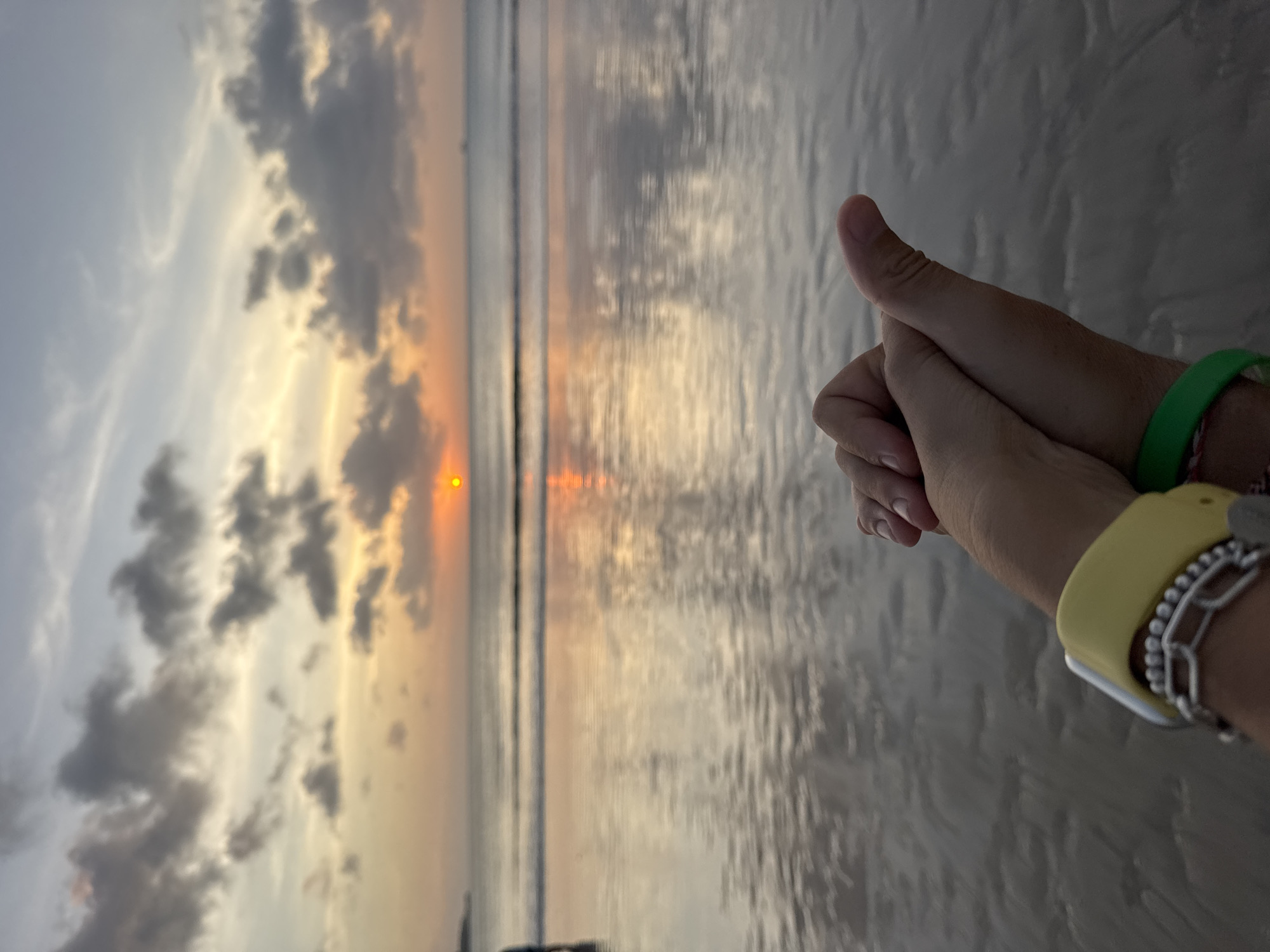A sunset view at Kuta Selatan beach, Indonesia, features two people holding hands in the foreground. The person on the right has a yellow watch band and a silver beaded bracelet on their wrist, while the person on the left wears a green wristband. The sun sets in the distance over the ocean, reflecting light on the water's surface. Clouds are scattered across the sky. The sandy beach is visible in the foreground.