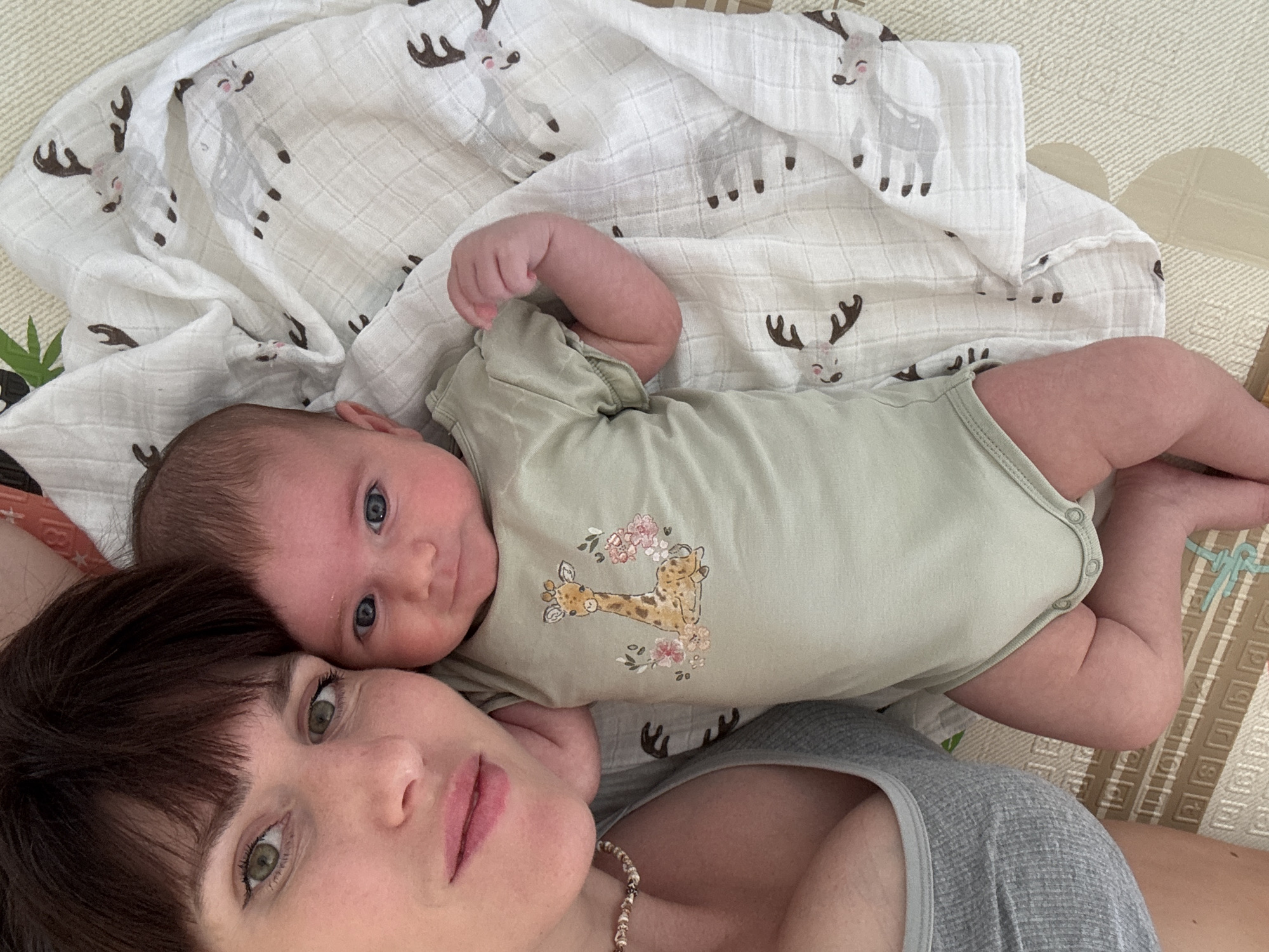 A woman and a baby are lying down, with the baby on top of the woman. They are on a light-colored, patterned blanket with illustrations of reindeer. The baby is wearing a pale green onesie with a giraffe and floral design. The woman has dark brown hair and bangs, and green eyes. The baby has blue eyes and dark hair. The scene appears to be indoors, possibly in a home, given the relaxed setting. The lighting suggests daytime. The overall mood is tender and intimate, capturing a moment between a mother and her child. There is no visible text in the image. The location context provided is Isola delle Femmine, Italy.