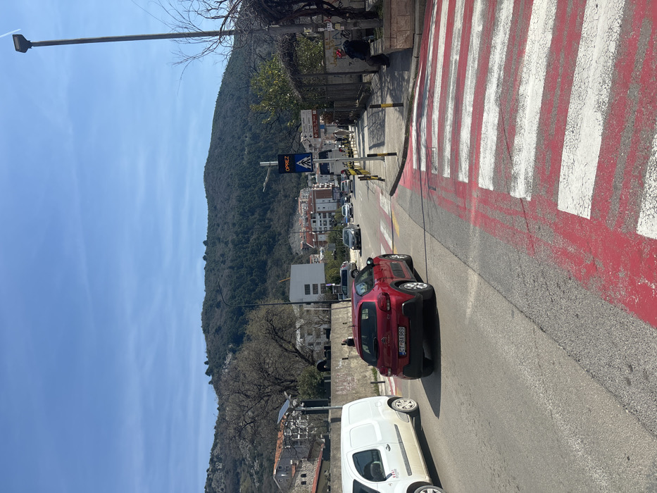 The image depicts a daytime street scene in Budva, Montenegro, specifically near the city center and Old Town, indicated by a directional sign. The view is along an asphalt road with a prominent red and white striped pedestrian crossing in the foreground. A red Citroën C3 Aircross, bearing Montenegrin license plate "CT BA 969," is positioned over the pedestrian crossing, facing away from the viewer. To the left, a white commercial van is partially visible, parked or stopped in the adjacent lane. Further down the road, additional vehicles, including cars and a bus, are in traffic.
On the right side of the road, a raised concrete sidewalk features a tall street light pole and yellow and black bollards. A person wearing a red hat and dark clothing is seated on a low stone wall, facing the road. Multiple pedestrians are visible walking along the sidewalk further down. A directional sign post on the right indicates "Budva centar" and "Stari grad" to the right. An overhead electronic sign displays "OPREZ" (Caution) above a blue pedestrian crossing symbol. On the left side of the road, behind a low stone wall, buildings and a bare tree are visible.
The background features numerous multi-story buildings, characteristic of an urban area, nestled against a steep, tree-covered mountain range under a clear blue sky with minimal cloud cover.