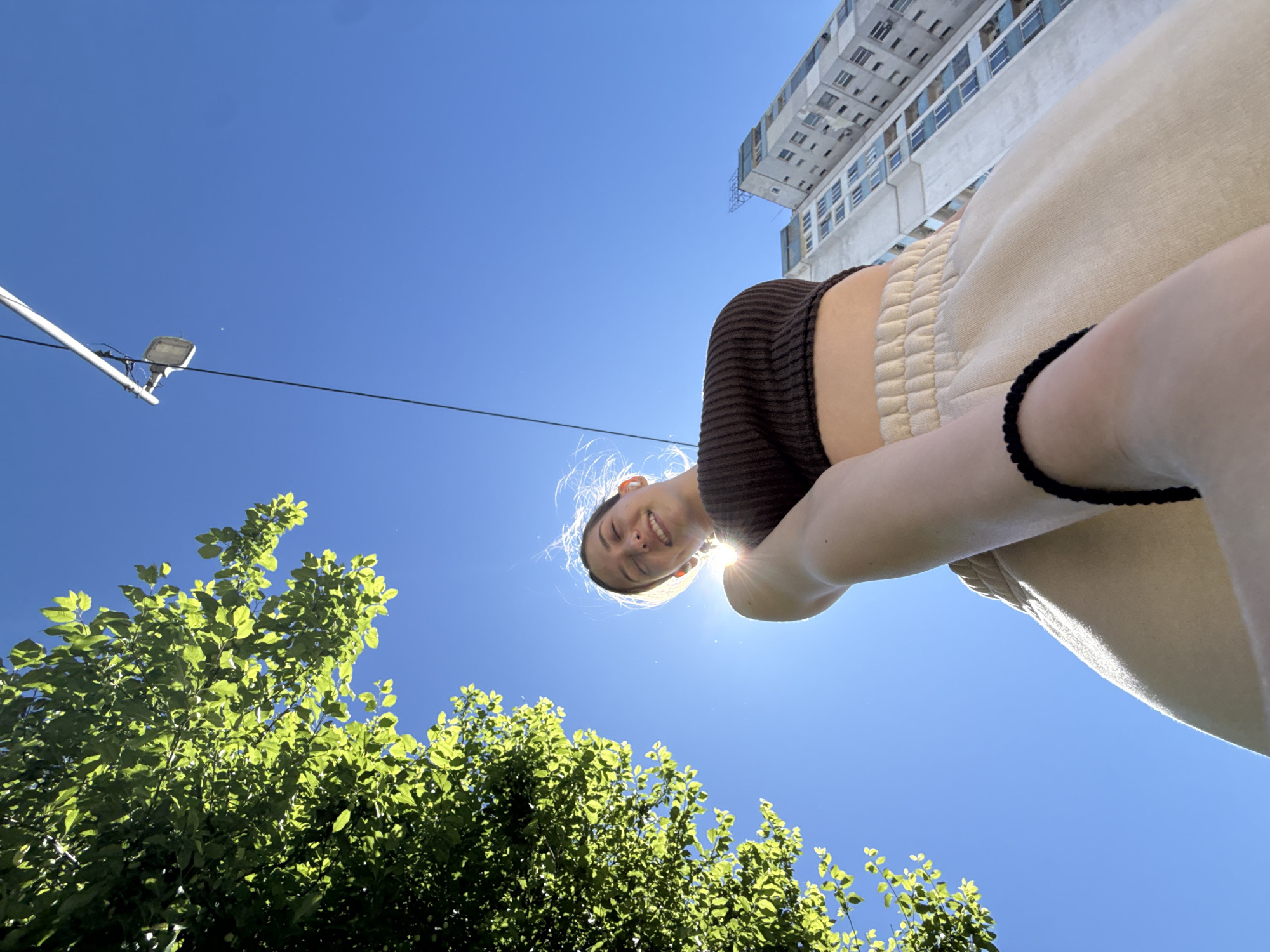 The image shows a person, presumably female, from a low-angle perspective. The person has fair skin, and blonde hair, and is wearing a dark brown top and tan pants with a black bracelet on her wrist. She is smiling. The background includes a clear, bright blue sky, green trees and a street lamp with power lines. A large, white multi-story building is visible at the top of the image. The location is most likely Sofia, Bulgaria.