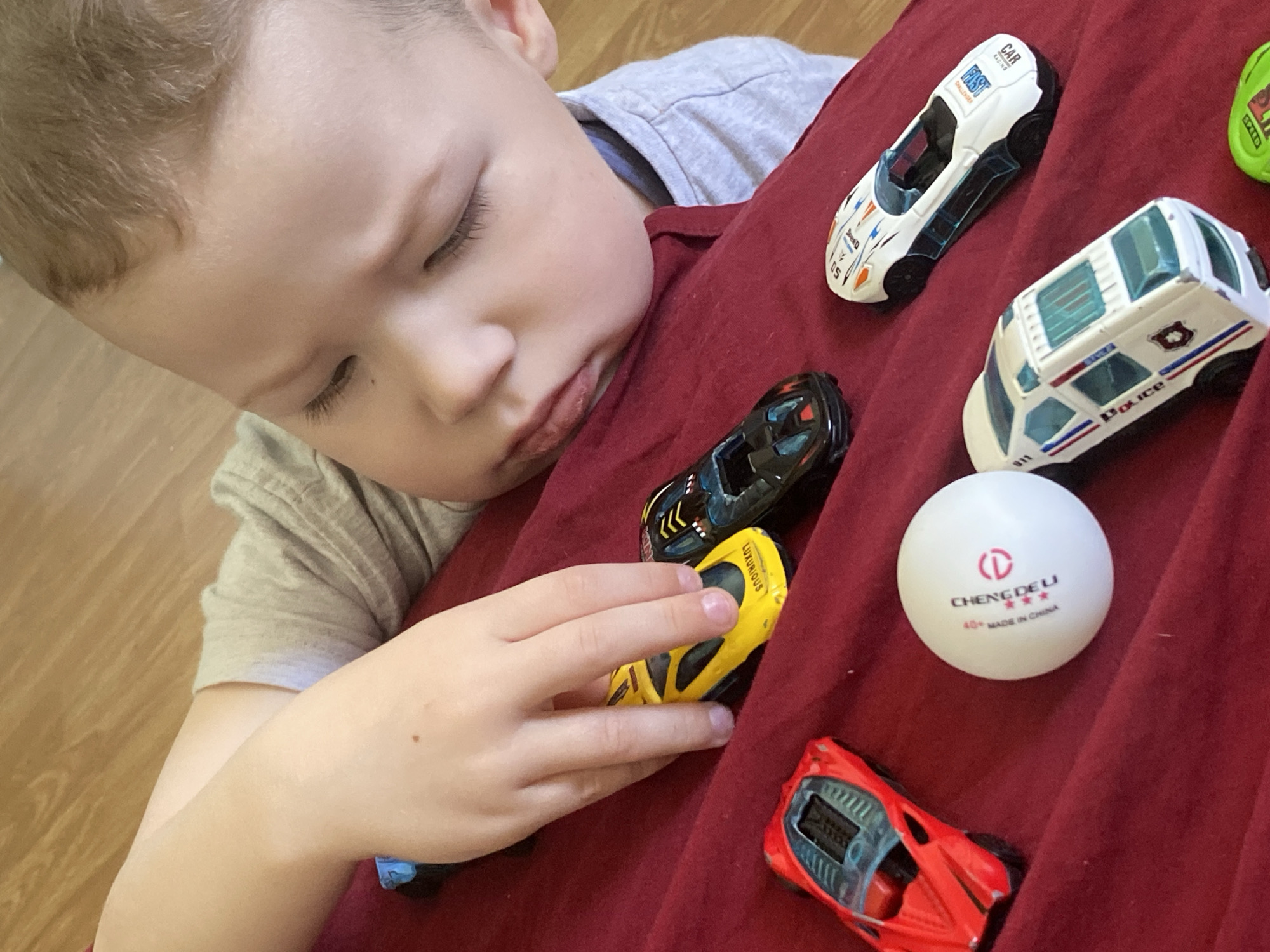 The image shows a young child lying on their side on a red fabric surface, deeply engaged with several toy cars and a ping-pong ball. The scene appears to be set indoors, possibly in a home in Sochi, Russia, with a wooden floor visible in the background.
The child, with light brown hair and fair skin, has their eyes downcast, suggesting focus or a moment of quiet contemplation. Their left hand gently rests on a yellow toy car, positioning it among the other toys. The expression on the child's face is serene and focused.
Around the child are several toy cars of various models: a white racing car, a black car, a white police car, and a red sports car. A green toy car is partially visible. A white ping-pong ball lies centrally among the toys.
Visible text includes:
* On the white racing car: "RACING", "CAR", "CHALLENGER", and "FAST".
* On the white police car: "POLICE" and "911".
* On the yellow toy car: "LUXURIOUS".
* On the partially visible green car: "SPEED".
* On the white ping-pong ball: "CHENG DELI", "40+", and "MADE IN CHINA".
The scene captures a moment of tranquil play and engagement from a child's perspective.