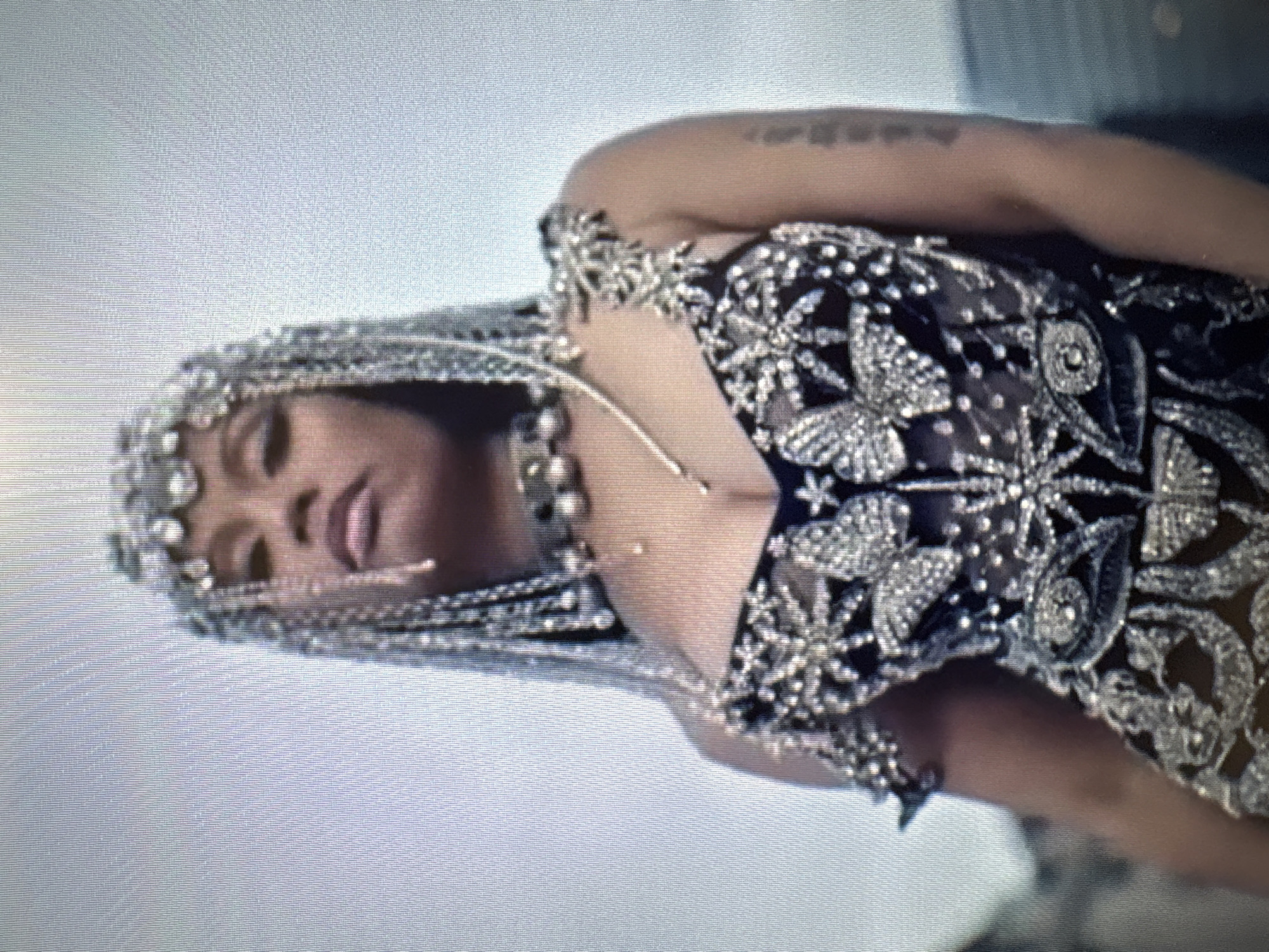 The image shows a close-up, waist-up shot of a woman. She is wearing an ornate, sparkling headdress with chains and jeweled embellishments, and a similarly decorated dark, sheer top with a sweetheart neckline. The top is adorned with butterflies and starburst patterns made of rhinestones or similar reflective material. A jeweled choker is visible around her neck, and delicate chains hang down from her headdress. Her left arm is visible, bearing tattoo markings. The background is a muted, slightly blurred sky, suggesting an outdoor setting. No specific details about the city of Sofia, Bulgaria, are visible in the image.