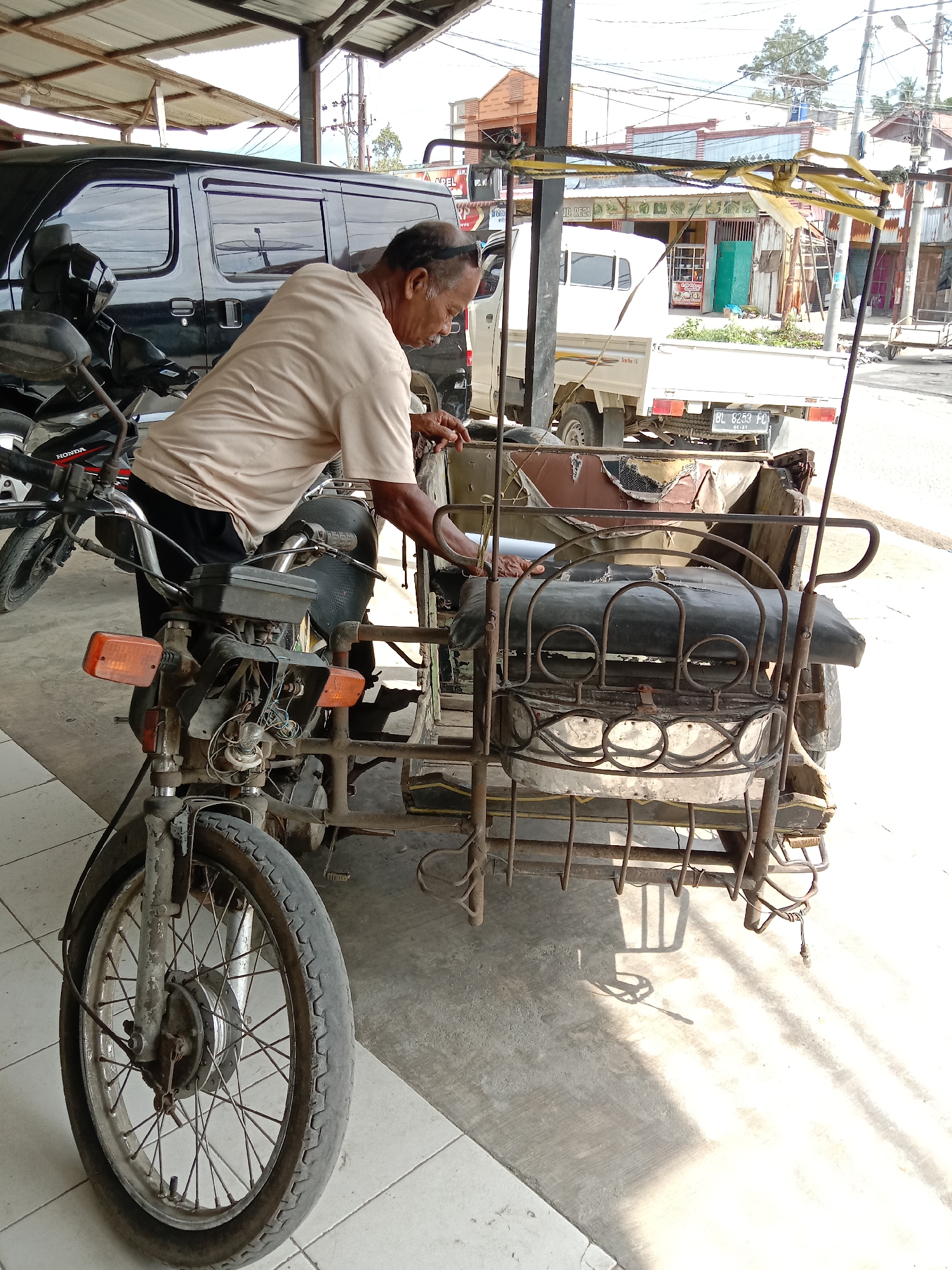 A man stands beside an older motorcycle and sidecar in Lhokseumawe, Indonesia. The motorcycle is facing the viewer with visible wires and an orange turn signal. The sidecar is made of metal tubes and has a seat. The man wears a tan shirt and dark pants. Behind the man, a black van is parked. In the background, there's a white truck and buildings. The scene is outdoors on a sunny day.