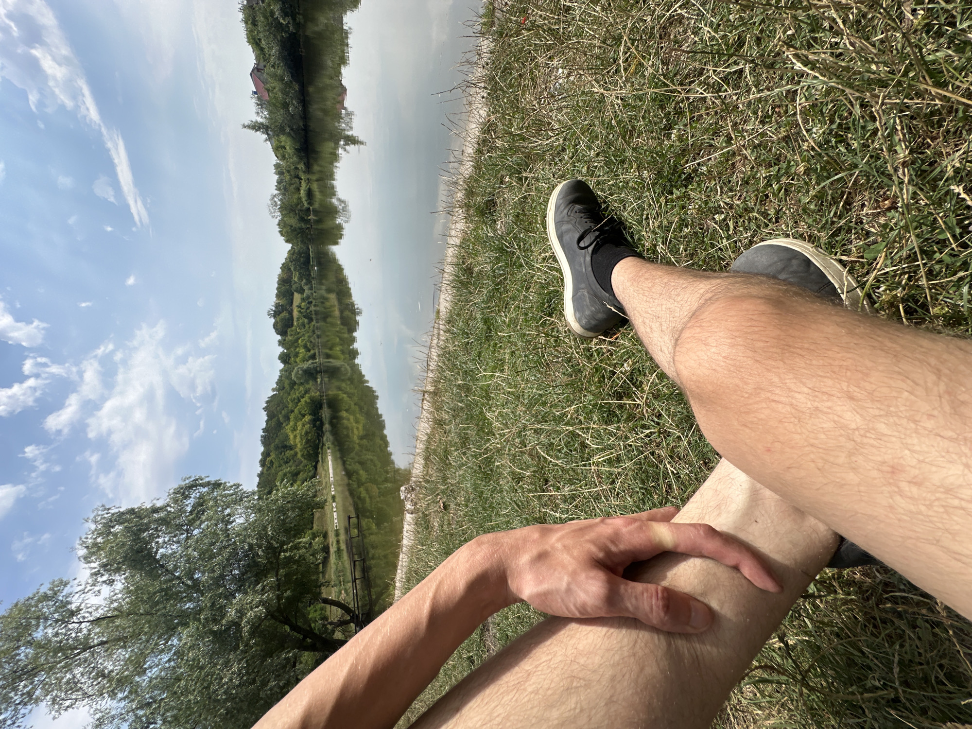 A first-person perspective shot shows a person sitting on a grassy bank next to a calm body of water. The person's legs and arm are visible in the foreground, with their hand resting on their shin. They are wearing dark shoes and black socks. The water reflects the sky and the trees on the opposite bank. The environment appears to be a park or natural area, possibly in Makiv, Ukraine, with lush green trees and a partly cloudy blue sky.