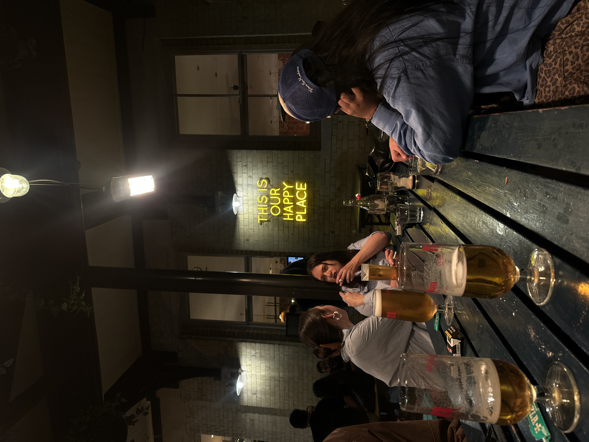 The image shows an outdoor seating area with a long, dark wooden table covered in glasses of beer. Three people are partially visible around the table. One person wears a blue shirt and a cap. Another is wearing a light blue striped shirt and has their hair up in a bun. The third person is also wearing a light blue shirt. At least three beer glasses are on the table, in various states of fullness. A neon sign on a brick wall behind the table reads "THIS IS OUR HAPPY PLACE." The setting appears to be nighttime. A flood light illuminates the seating area. Windows are visible on the wall behind the neon sign. The city is London, United Kingdom.