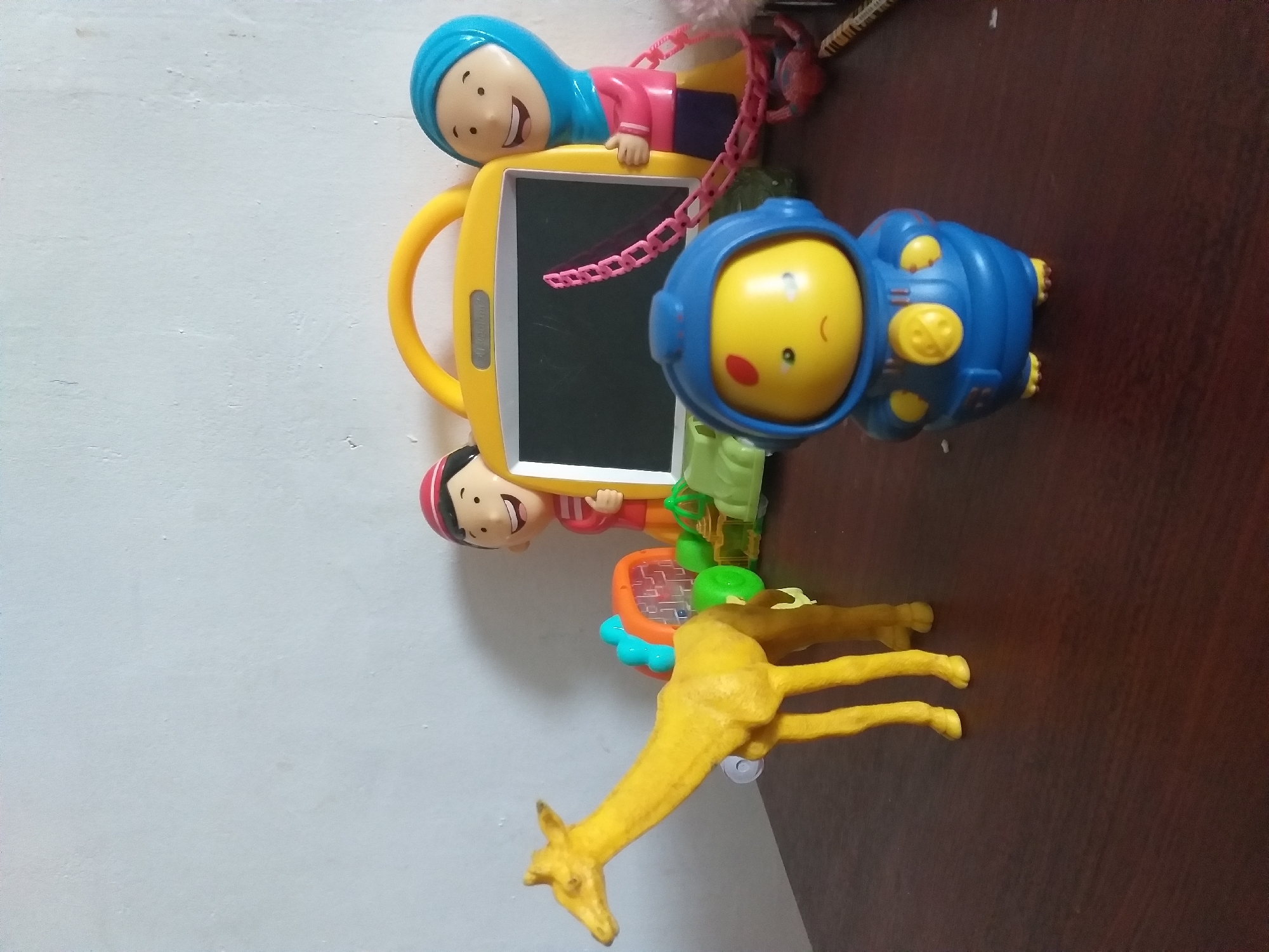 An indoor still life features several children's toys arranged on a dark wooden surface against a textured white wall. On the left, a bright yellow toy giraffe stands with its head turned to the left, its body slightly bent. To the right of the giraffe, a green toy vehicle with blue and orange accents is visible. In the center background, a yellow toy drawing board with a handle is propped up. A cartoonish figure of a boy wearing a red cap and striped shirt stands behind the drawing board on the left. To the right of the drawing board, a cartoonish figure of a girl wearing a blue hijab and pink shirt stands, giving a thumbs-up. In the foreground, a blue and yellow toy astronaut figure with a sad expression is positioned centrally. A pink chain dangles from the drawing board, connecting to the astronaut. The scene is dimly lit, casting shadows on the wall and surface. The location is the city of Air Jamban, Indonesia.