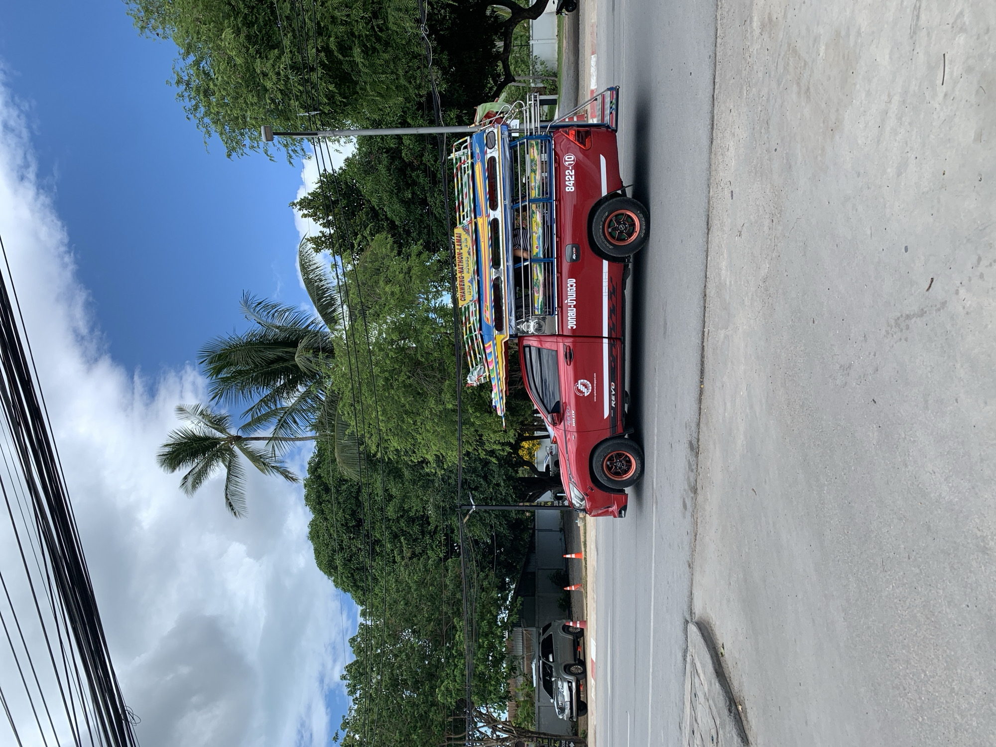 A red Toyota Hilux Revo pick-up truck, customized as a songthaew (a passenger vehicle with a canopy and benches), is captured in motion on an asphalt road in Baan Bo Phut, Thailand. The truck features a brightly colored, multi-tiered canopy structure on its bed, adorned with yellow, blue, red, and green accents. A sign on the front of the canopy displays "CHAWENG-NATHON-LAMAI" in English and Thai script, indicating its route. The truck's side bears the text "REVO HILUX," "WWW.UPKOHSAMUI.BLOGSPOT.COM," and "อวนบ้านเฉวง" (Ao Wan Ban Chaweng), along with a white logo. The vehicle's wheels have distinctive orange-red rims. A rear metal ladder structure is visible, leading to the passenger area, where a person in a striped shirt is partially visible. The truck's rear side displays the number "8422-10."
To the left of the road, a silver SUV, resembling a Toyota Fortuner, is parked on a dirt shoulder, with two orange traffic cones positioned nearby. The road itself is gray asphalt, bordered by a concrete curb and a sidewalk on the left. The background is dominated by lush green foliage, including numerous tropical trees and tall palm trees, indicative of a tropical environment. Overhead, a dense cluster of black utility lines runs horizontally across the top of the frame. The sky above is partially cloudy, showing patches of clear blue interspersed with white clouds. A dark gray utility pole with a streetlight is visible on the right side of the road, partially obscured by trees. Fences and vague building outlines are present in the distant background.