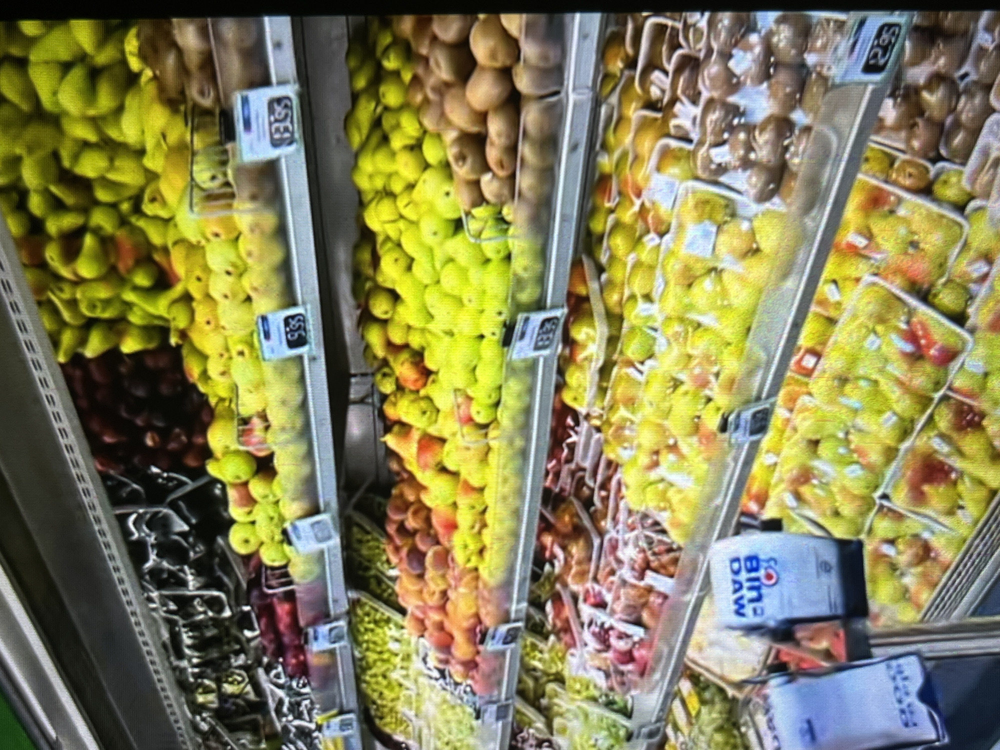 The image displays a grocery store produce section, specifically focusing on shelves stocked with various fruits. The arrangement suggests an indoor market or supermarket environment, with multiple tiers of shelving. The fruits are presented in bulk and in pre-packaged containers, with some items featuring price tags. The colors are vibrant, indicating fresh produce. The presence of a shopping cart and other shoppers is not evident in this specific frame. The lighting appears to be artificial, typical of an indoor retail setting.
This scene is located in the city of Tinaquillo, Venezuela, within a grocery store's produce aisle. The shelves are filled with an assortment of fruits, including apples, pears, peaches, and kiwis. Some of the fruits are loose, while others are in transparent plastic packaging. Price tags are visible on some of the items, indicating costs such as "9.95" and "13.95" (currency not specified). In the foreground, a blue and white dispenser with writing in Spanish, likely for plastic bags or labels, is present. The overall impression is one of a well-stocked and organized produce section in a Venezuelan supermarket.