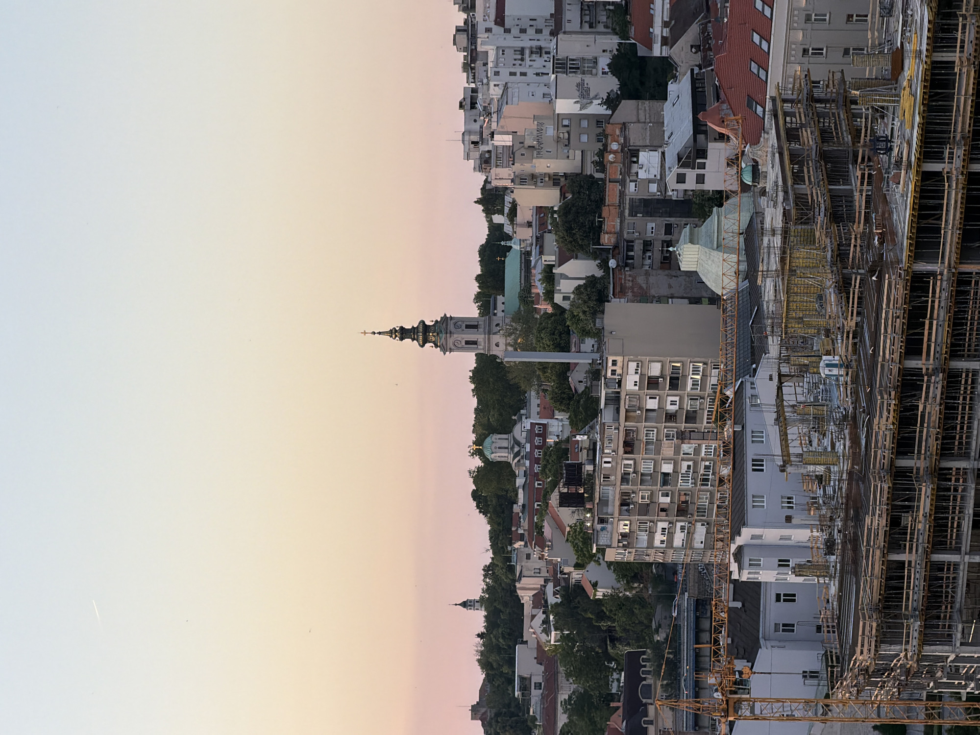 The image depicts a cityscape with a clear sky transitioning from light to dark from left to right, suggesting either sunrise or sunset. The urban landscape consists of buildings of varying heights and architectural styles. A prominent church steeple rises above the tree line in the middle of the frame, possibly representing the Serbian Orthodox Church. A construction site with scaffolding and cranes is visible on the right side of the image, indicating ongoing urban development. The buildings range from older structures to modern apartment blocks. The trees add a natural element to the cityscape. The scene is most likely Belgrade, Serbia, judging by the architectural styles and urban development patterns visible in the image.