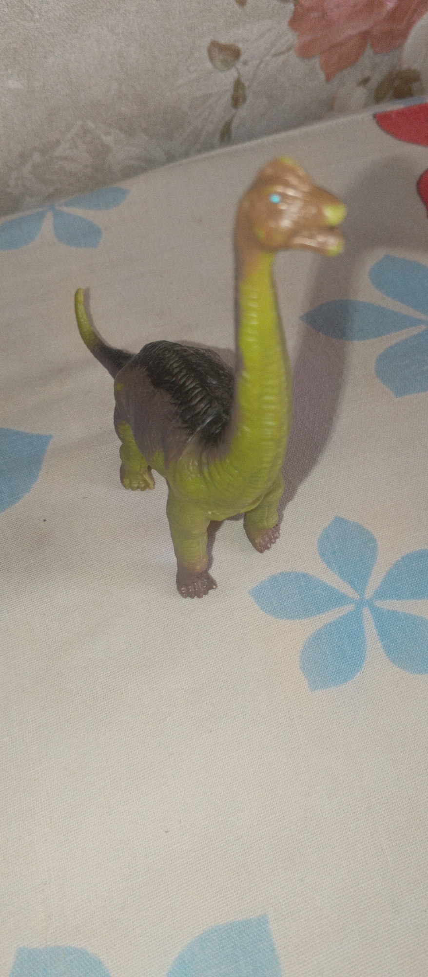 A plastic toy dinosaur, colored green and brown with blue eyes, stands on a white fabric surface patterned with light blue flowers. The dinosaur's head is tilted slightly to the right, and its long neck is curved. The background consists of a textured surface, possibly wallpaper, with floral patterns in muted tones. The lighting suggests an indoor environment. The scene is static and devoid of human presence.