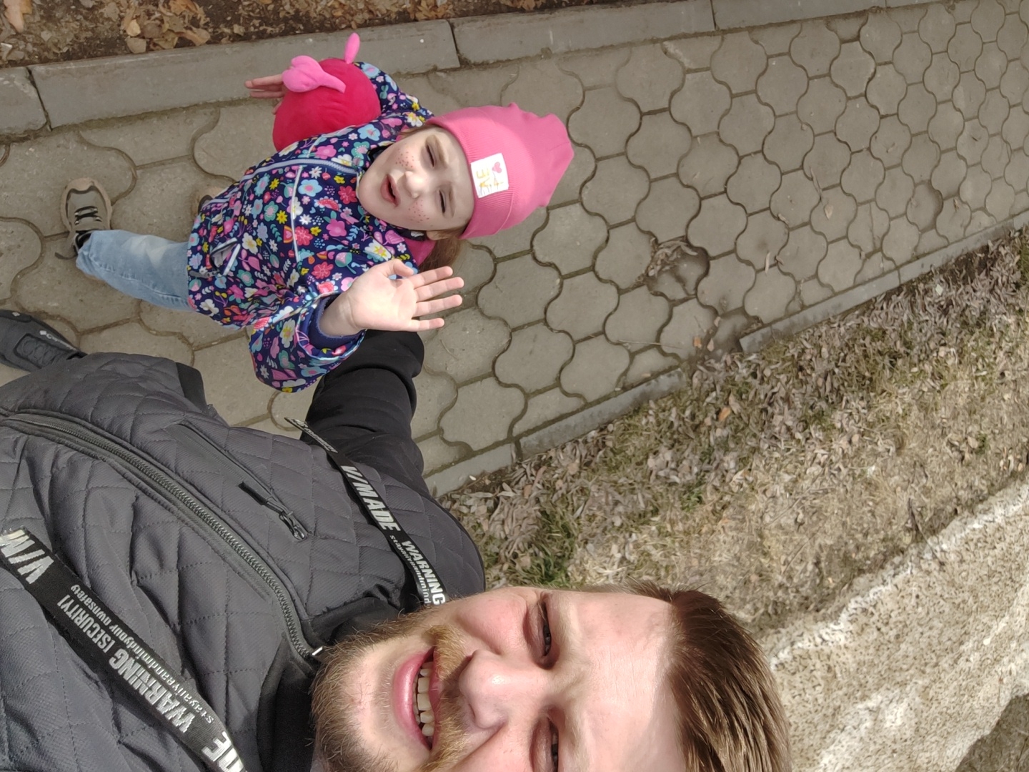 A slightly canted, medium-shot, taken from a low angle, showcases a man and a young girl on a city sidewalk. The sidewalk is made of gray interlocking pavement stones. The man's face, fair-skinned with a brown beard, occupies the bottom portion of the frame, with a smiling expression and teeth visible. He wears a dark gray quilted jacket with a black sleeve. White text on black straps is visible around his collar. The young girl is above him, wearing a pink knit hat, a patterned jacket, and jeans. She is looking at the camera and waving, and appears to be Caucasian. The background shows a patch of dead grass and dead leaves and twigs, along the edge of the sidewalk, and a partial patch of white. The location is identified as the city of Unknown, Unknown.