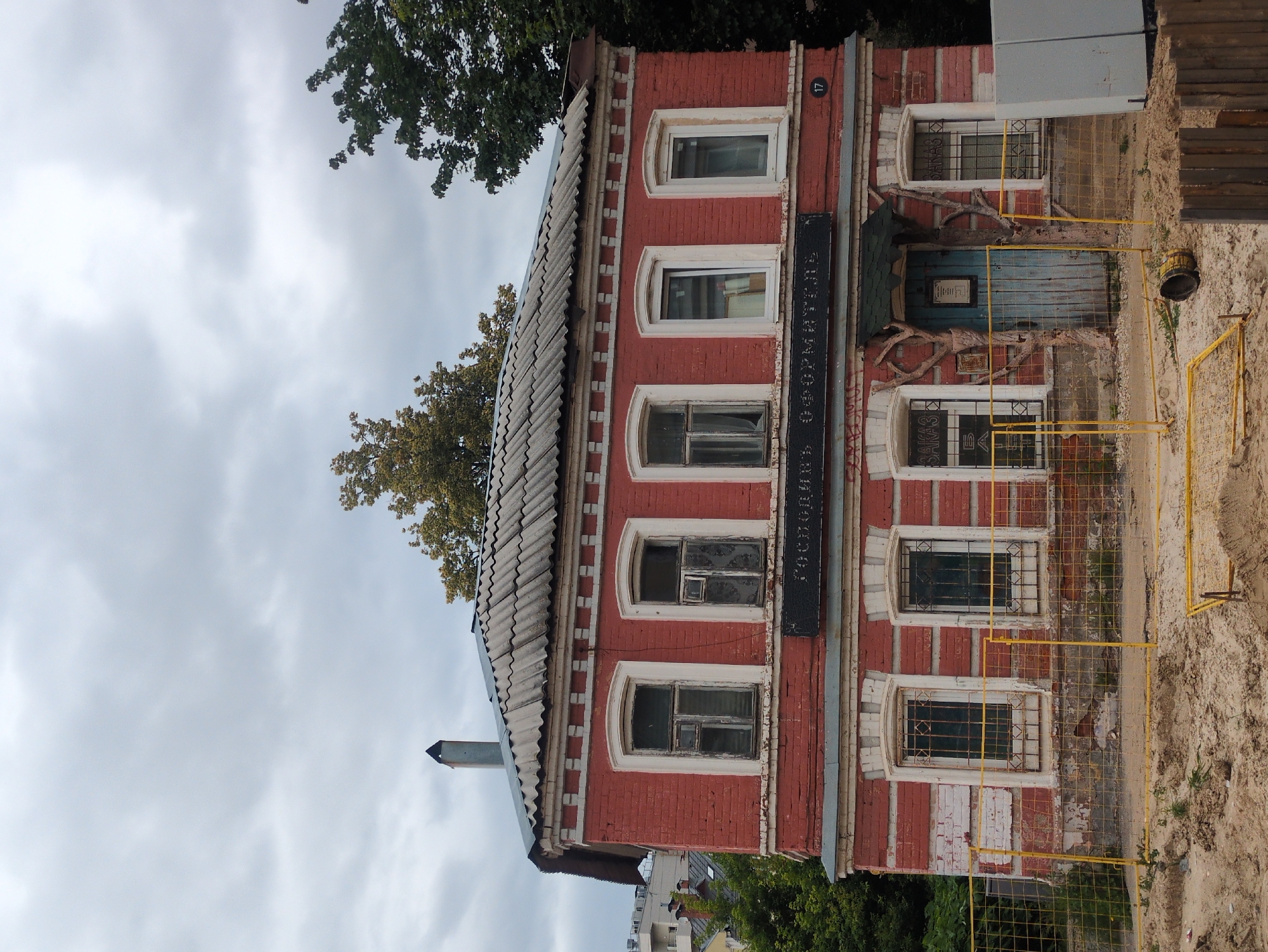 The image shows a two-story red brick building in Nizhny Novgorod, Russia. The building has a corrugated metal roof and several windows with white trim. Above the entrance, a sign reads "Господинь Оформитепь". A blue wooden door is located in the center of the building, framed by tree branches. A yellow fence surrounds the front of the building, and the ground is covered in sand and dirt, suggesting ongoing construction or renovation. Trees are visible in the background, and the sky is overcast.