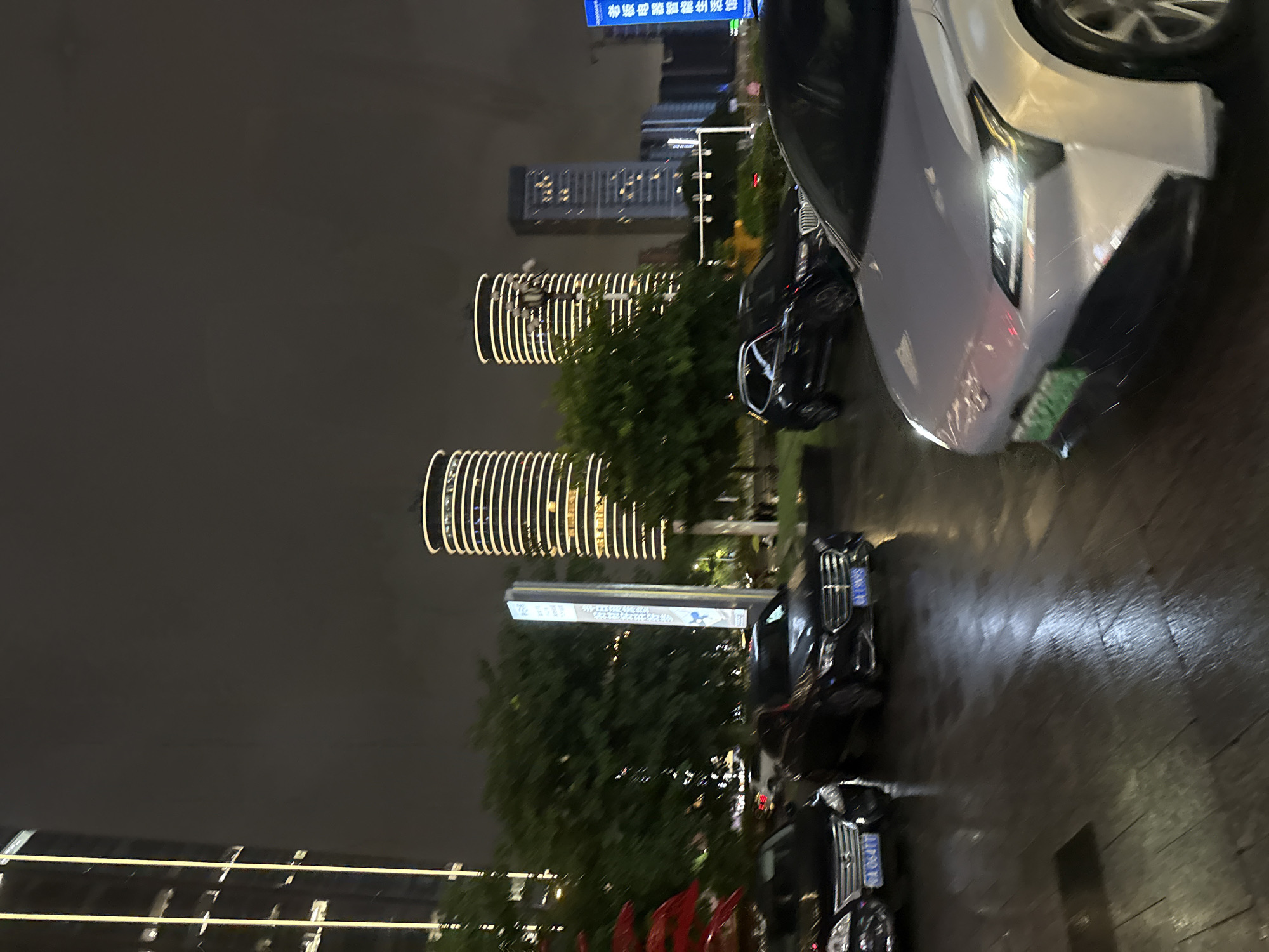 In the Tianhe District of China, at night, a view of a wet, paved street shows several cars. In the foreground, a white electric vehicle is parked facing right, its headlights on, revealing a license plate starting with "93". To the left, two black sedans are also parked. The car closest to the viewer has a license plate starting with "GX 06477", and the car behind it has a license plate beginning with "GX 19NVS". In the background, two cylindrical skyscrapers with illuminated horizontal bands rise into the dark sky, along with other urban buildings. Green trees are visible between the cars and the skyscrapers, and a tall, slender signpost stands near the trees. The ground appears wet, reflecting the ambient light, suggesting recent rain.