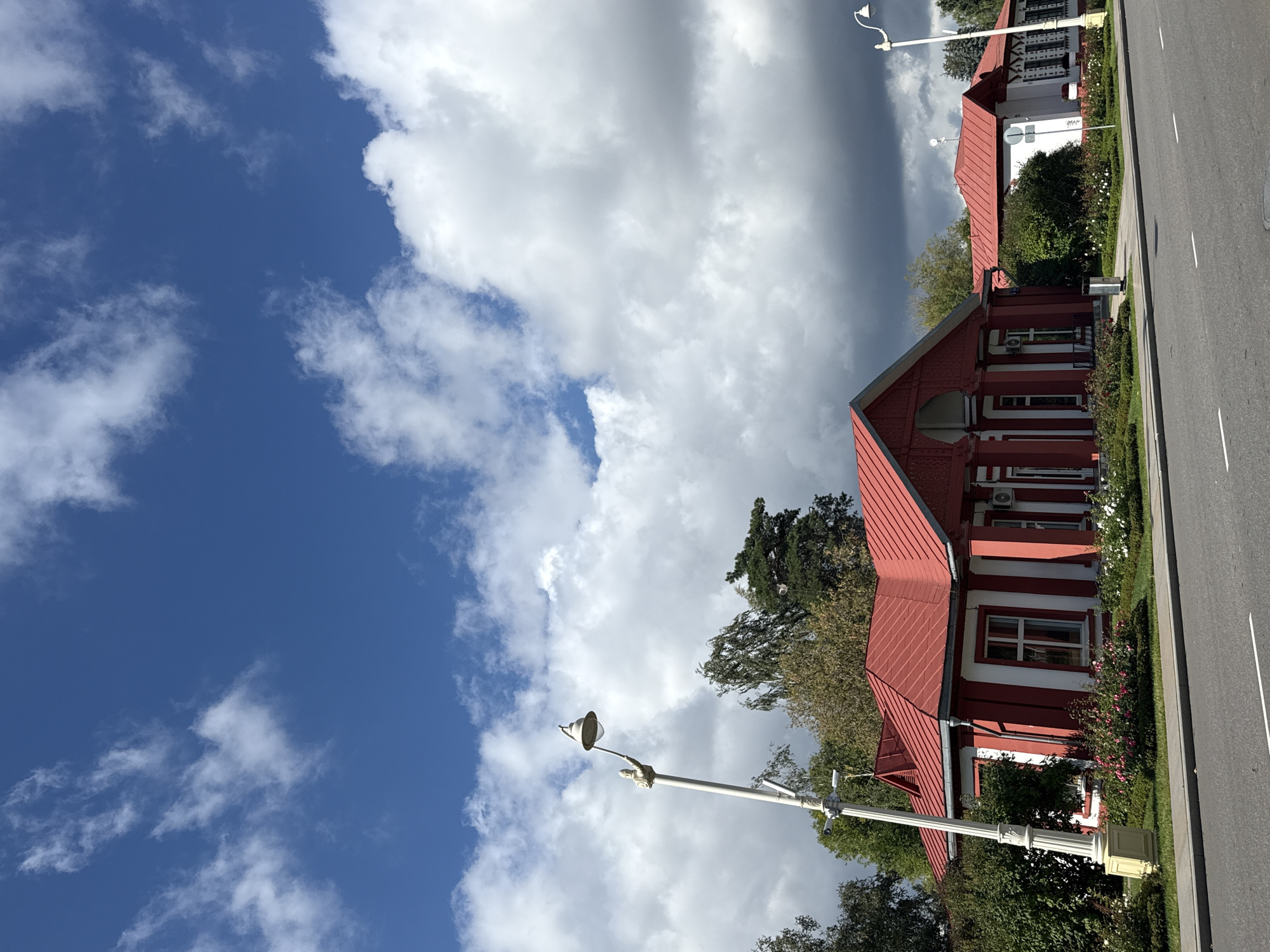 The image shows a red-roofed building with white walls and red trim. The building has several windows and columns. There is a lamppost in front of the building, and a street in the foreground. The sky is blue with some white clouds. The scene is located in Moscow, Russia.