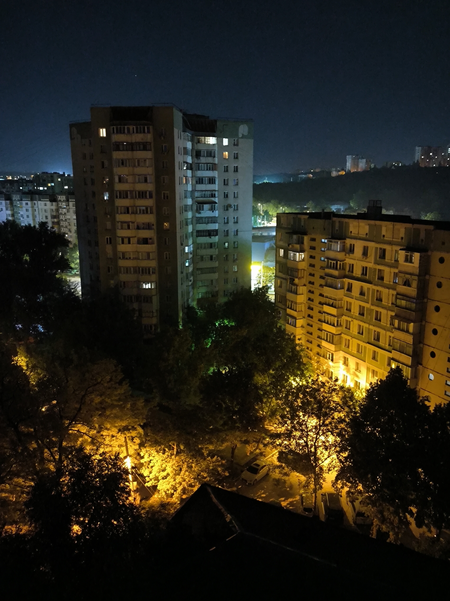 This is a nighttime, high-angle shot of a residential area in Chișinău, Moldova. The scene is dominated by several tall apartment buildings, some of which are illuminated with interior lights, suggesting occupancy. The architecture appears to be typical of Soviet-era mass housing, characterized by concrete structures and repetitive window patterns.
In the foreground, several trees are visible, their leaves illuminated by artificial lights, creating a dappled effect. The ground level reveals a parking area with several parked cars, their forms partially obscured by shadows and the surrounding foliage. The lighting in the scene is predominantly artificial, casting a warm glow from streetlights and building interiors onto the urban landscape.
In the background, more apartment buildings and distant city lights are visible, indicating the density of the urban environment. The sky is dark, characteristic of nighttime, with a faint haze or light pollution visible on the horizon. The overall impression is of a quiet, residential district at night, with the built environment and natural elements coexisting under the artificial illumination.