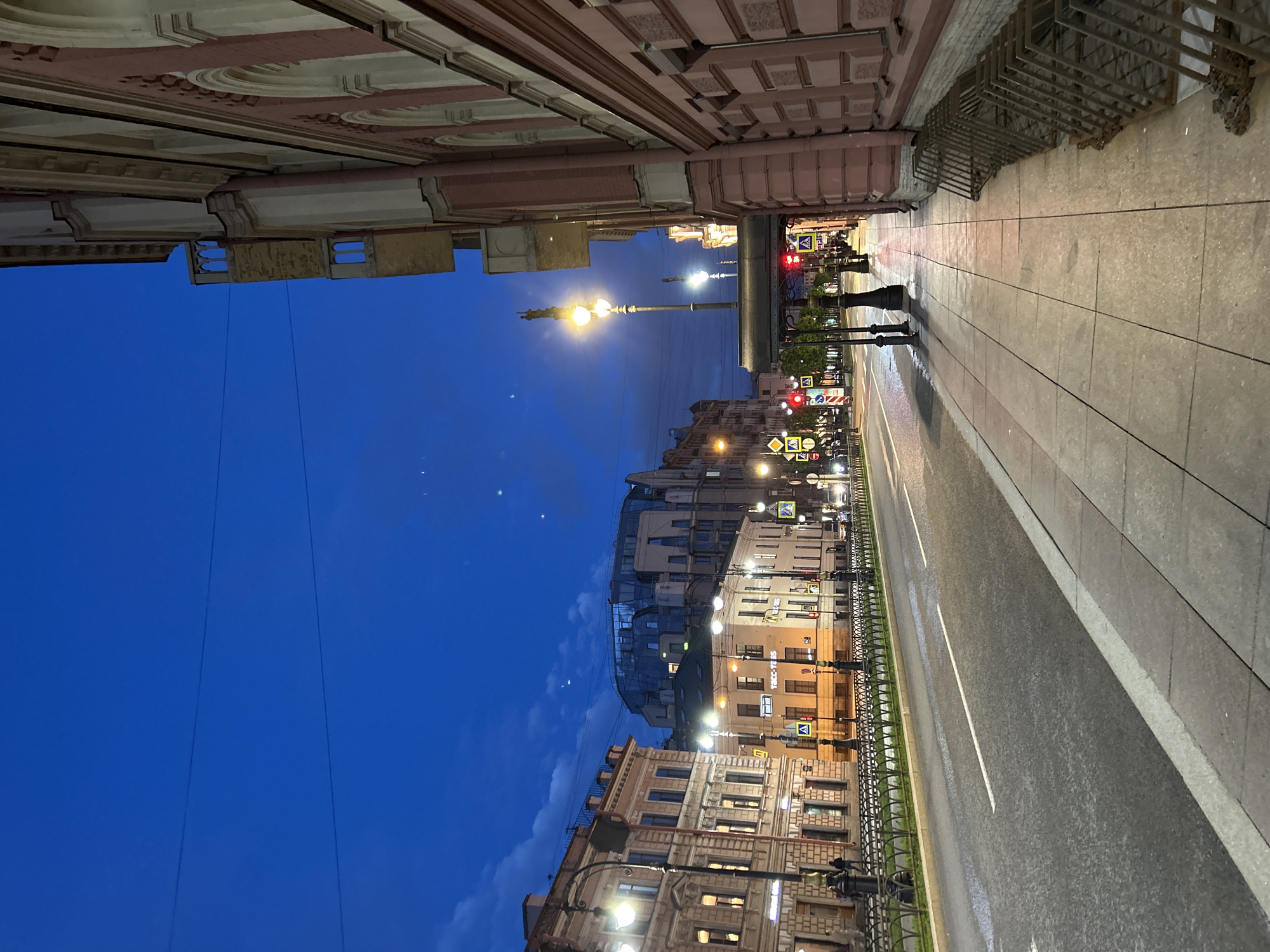 The image depicts a street scene at dusk in a city. The sky is a deep blue with some scattered clouds. A road with white lane markings runs through the center of the frame, reflecting ambient light. Sidewalks flank the road, with one side featuring a row of metal structures that appear to be bicycle racks. Buildings with architectural details line the street, some illuminated with interior lights. Streetlights and traffic signals are visible, indicating active urban infrastructure. A few people are seen walking on the sidewalk in the distance, and vehicles are present on the road. The overall impression is of a European city at twilight, possibly during the summer due to the brightness of the sky.