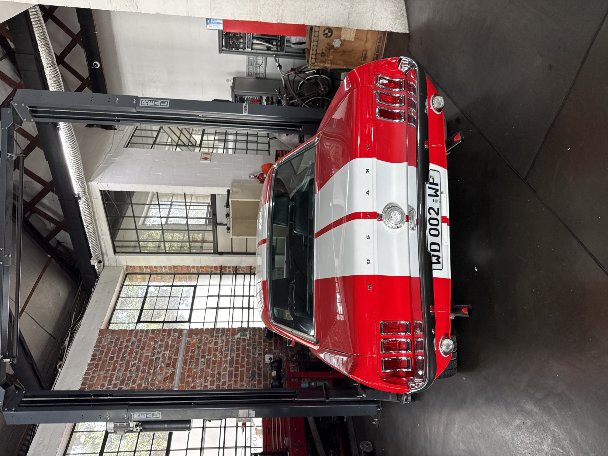 An overhead view captures a striking red and white vintage Ford Mustang coupe parked on a car lift inside a spacious workshop. The car features a prominent white racing stripe running down its center, contrasting with the vibrant red body. "MUSTANG" lettering is visible on the trunk lid, along with a circular emblem. The license plate "WD 002 WP" is clearly displayed, indicating a South African registration (consistent with Cape Town context). Chrome accents adorn the taillights and bumper.
The setting is an industrial-style garage or workshop, characterized by exposed brick walls, large grid-patterned windows allowing natural light, and visible ceiling structures including beams and ventilation ducts. The dark grey car lift, prominently positioned, bears the brand name "REAL" on its side. In the background, various workshop equipment, tools, shelves, and a bicycle are visible, alongside a cardboard box marked "BMW" and a small red fire extinguisher sign with an arrow. The floor is a dark, possibly concrete or epoxy, surface. A security camera is mounted high on a wall. The car appears to be either undergoing maintenance or stored on the lift.