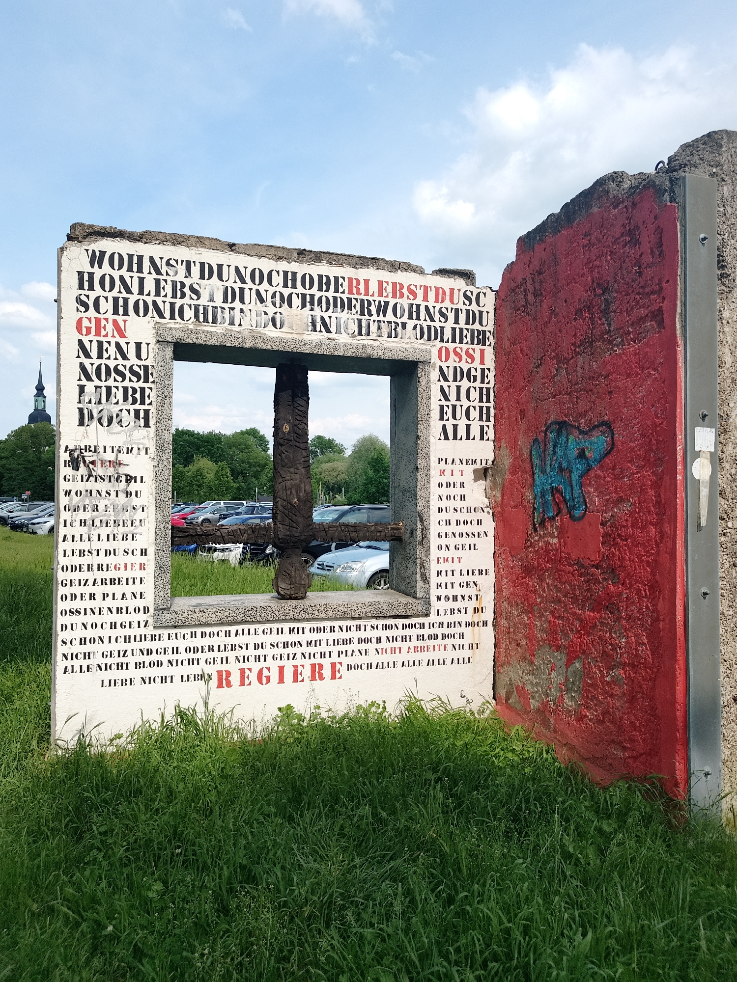 The image shows a concrete structure in a grassy area, with cars and greenery visible in the background, suggestive of a public park or parking area adjacent to a green space. The structure is divided into two main sections: a white, rectangular frame filled with German text and a separate, red-painted panel to the right, adorned with a blue graffiti design. The text on the white frame consists of various words and phrases, with some highlighted in red. A wooden post or carved element is centrally placed within the opening of the white frame. The right panel is textured and appears weathered. The overall scene suggests a contemporary art installation or memorial.
