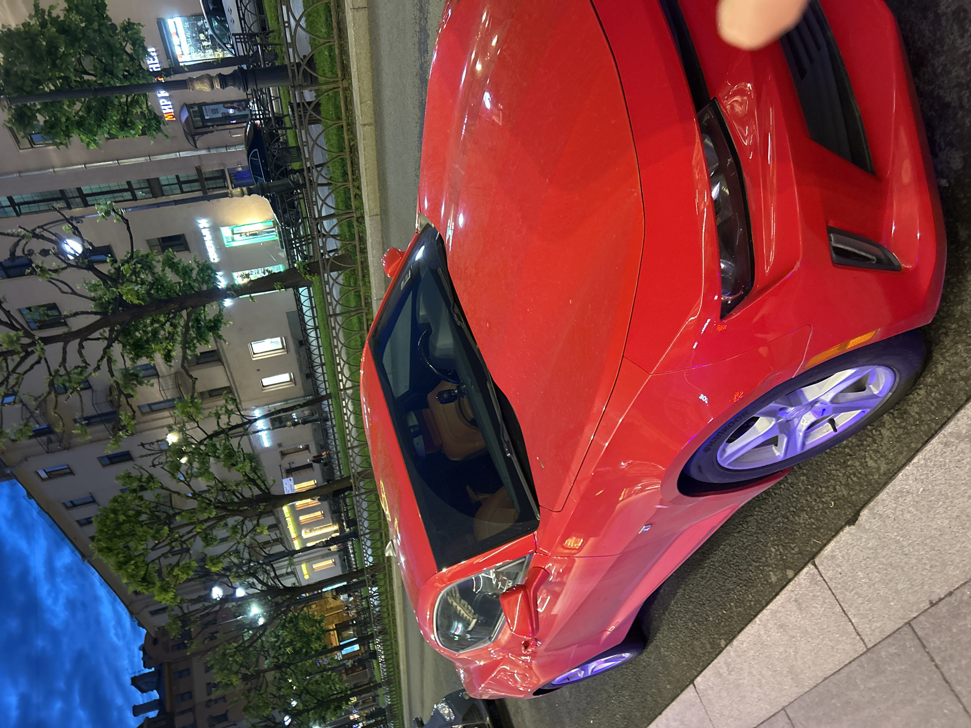 A bright red Chevrolet Camaro is parked on the side of a street in an urban setting, possibly at dusk or night given the dark blue sky and lit buildings. The car is positioned at an angle, with its front portion closest to the viewer. The car has a sporty design, with sharp lines and a low profile. The interior of the car, visible through the windows, appears to have tan or brown leather seats.
The street is lined with trees and ornate streetlights. In the background, there are multi-story buildings with illuminated windows and some storefronts. A wrought-iron fence separates the sidewalk from a grassy area in front of the buildings. The sidewalk itself is paved with light-colored stones and concrete. Another dark-colored vehicle is partially visible behind the Camaro on the left side of the image. The overall impression is that of a modern car parked on a historic city street.