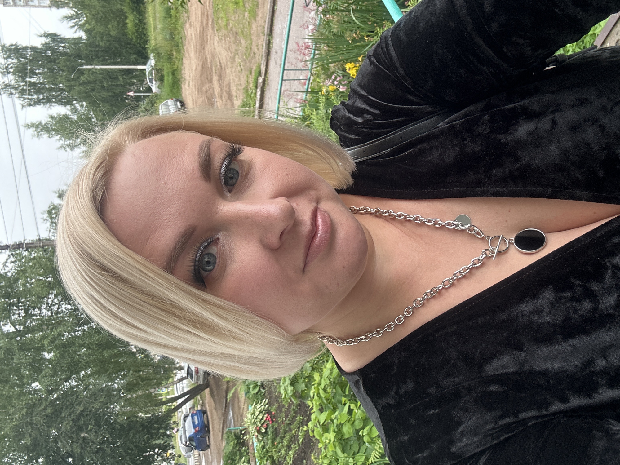 A woman with blonde hair and blue eyes is photographed outdoors in Chaikovsky, Russia. She is wearing a black velvet top and a silver chain necklace with a black pendant. The background shows a street with parked cars and trees. The sky is overcast.