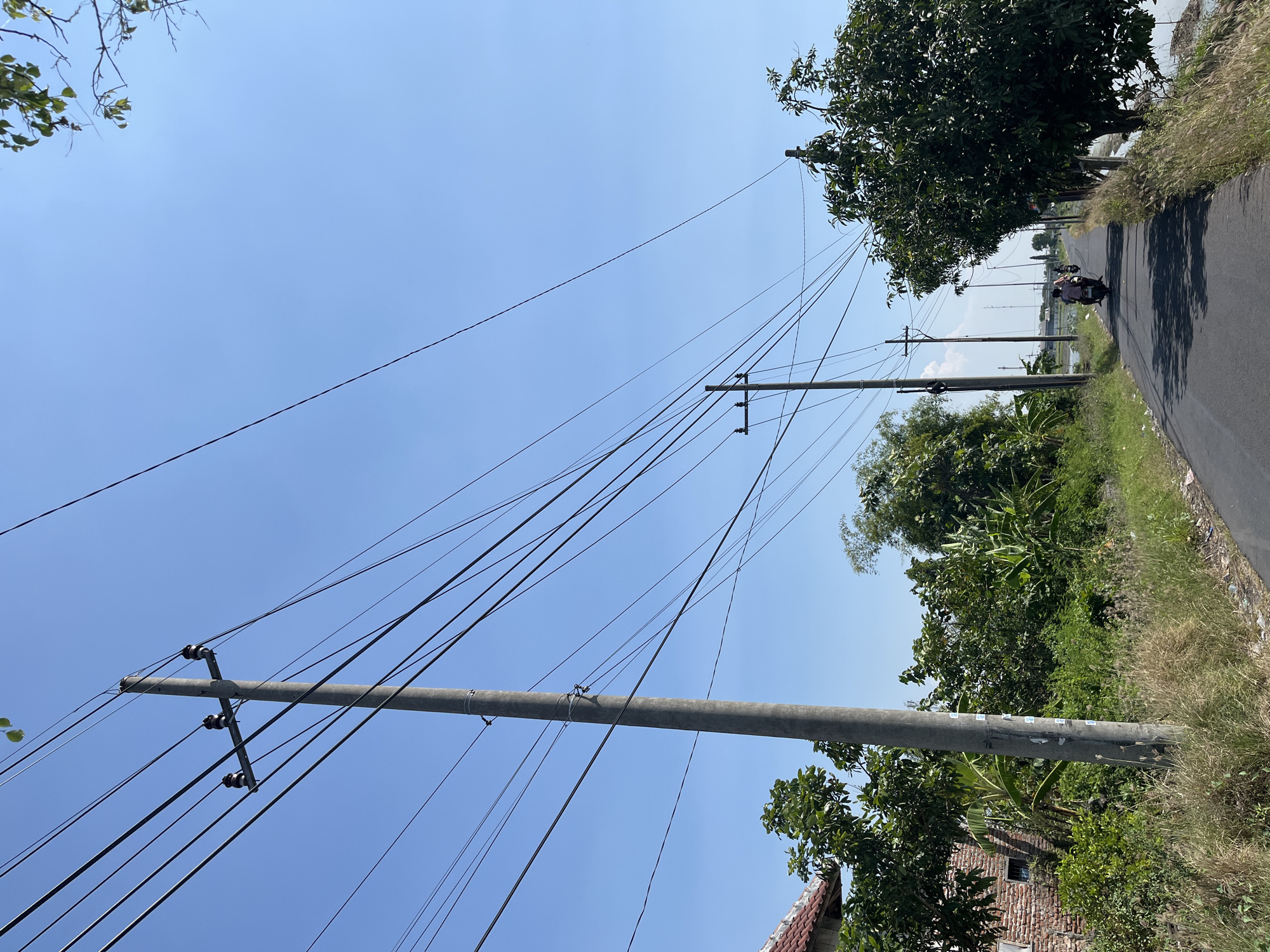 A paved road curves into the distance under a clear blue sky in Pesawahan, Indonesia. Tall concrete utility poles with numerous wires stretch across the scene, lining the road and disappearing into the horizon. To the left of the road, a small brick building with a red-tiled roof peeks through dense green foliage, including trees and what appear to be banana plants. The right side of the road is bordered by tall grasses and the edge of a body of water, likely rice paddies. Two people are riding a motorcycle down the road, casting shadows on the asphalt. Further down the road, two more motorcycles are visible, also carrying passengers. The overall scene depicts a rural or semi-rural environment in Indonesia on a sunny day.