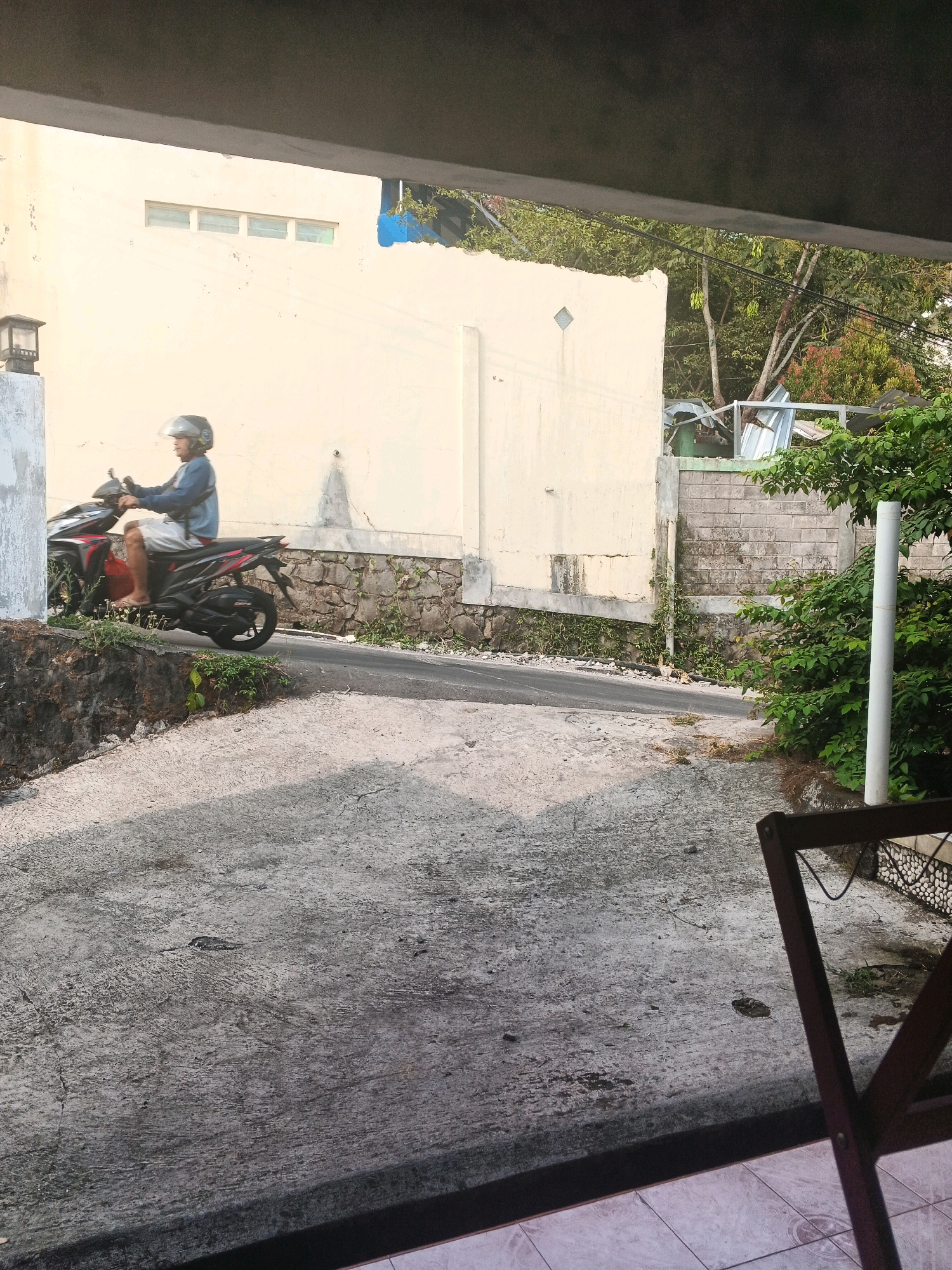 A person wearing a blue shirt and a white helmet rides a red and black motorcycle on a paved road. The road is bordered by a stone wall and overgrown vegetation on the left, and a cream-colored building on the right. The building has a row of small windows near the top. In the background, there are trees, power lines, and a partially collapsed metal structure. The foreground shows a concrete surface and a wooden chair. The scene appears to be a residential area in Keji, Indonesia, under natural daylight.