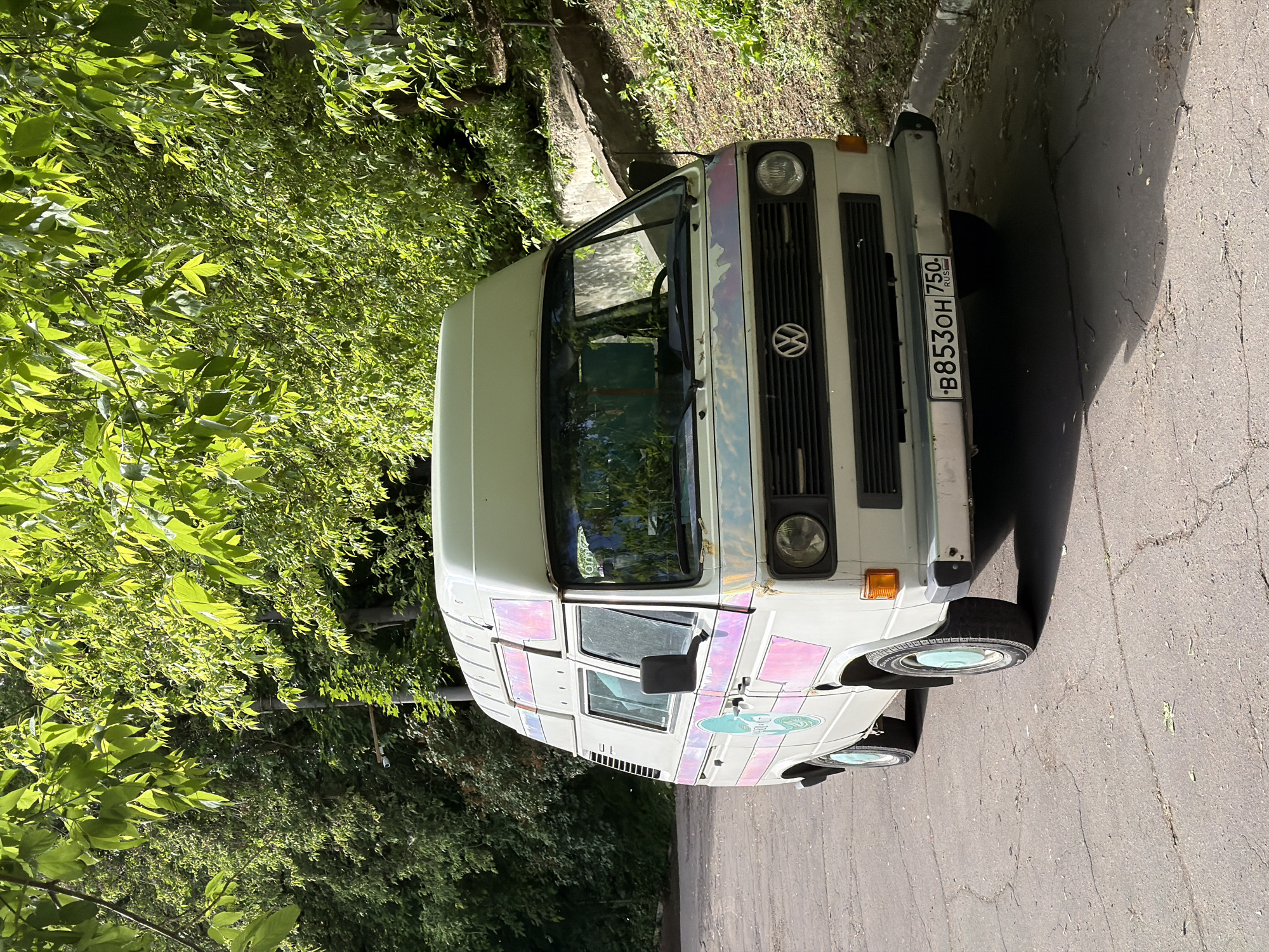 A white Volkswagen van with a Russian license plate "B853OH 750" is parked on asphalt near lush green foliage, possibly in Moscow, Russia, as indicated by the license plate. The van has decorative elements on its side. The background consists of trees and greenery suggesting a park or residential area. The photograph is taken during daylight.