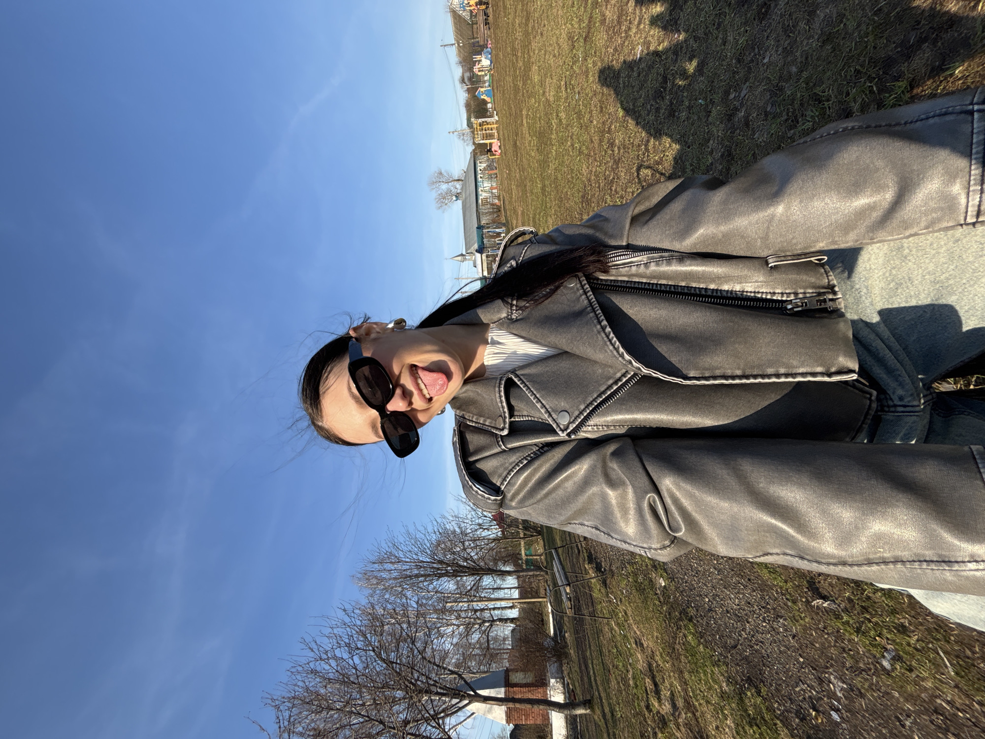 The image shows a woman in sunglasses and a leather jacket taking a selfie, sticking her tongue out. The background consists of a clear blue sky, trees, grass, and buildings, and possibly a playground area. The picture was taken in the city of Фоки, Russia.