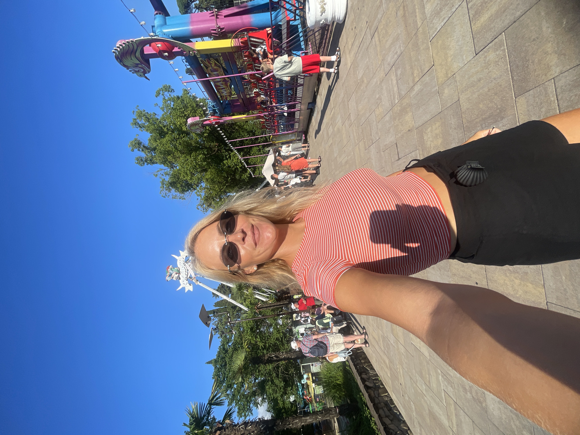 A blonde woman with sunglasses and a red and white striped crop top takes a selfie outdoors on a sunny day. She is standing on a paved walkway in what appears to be an amusement park. Behind her, a brightly colored amusement ride with a dragon-like structure is visible, as are other people walking and enjoying the park. Trees and palm trees provide greenery, and the sky is a clear blue. The scene is consistent with an outdoor recreation area in Sochi, Russia, a city known for its entertainment venues and attractions.