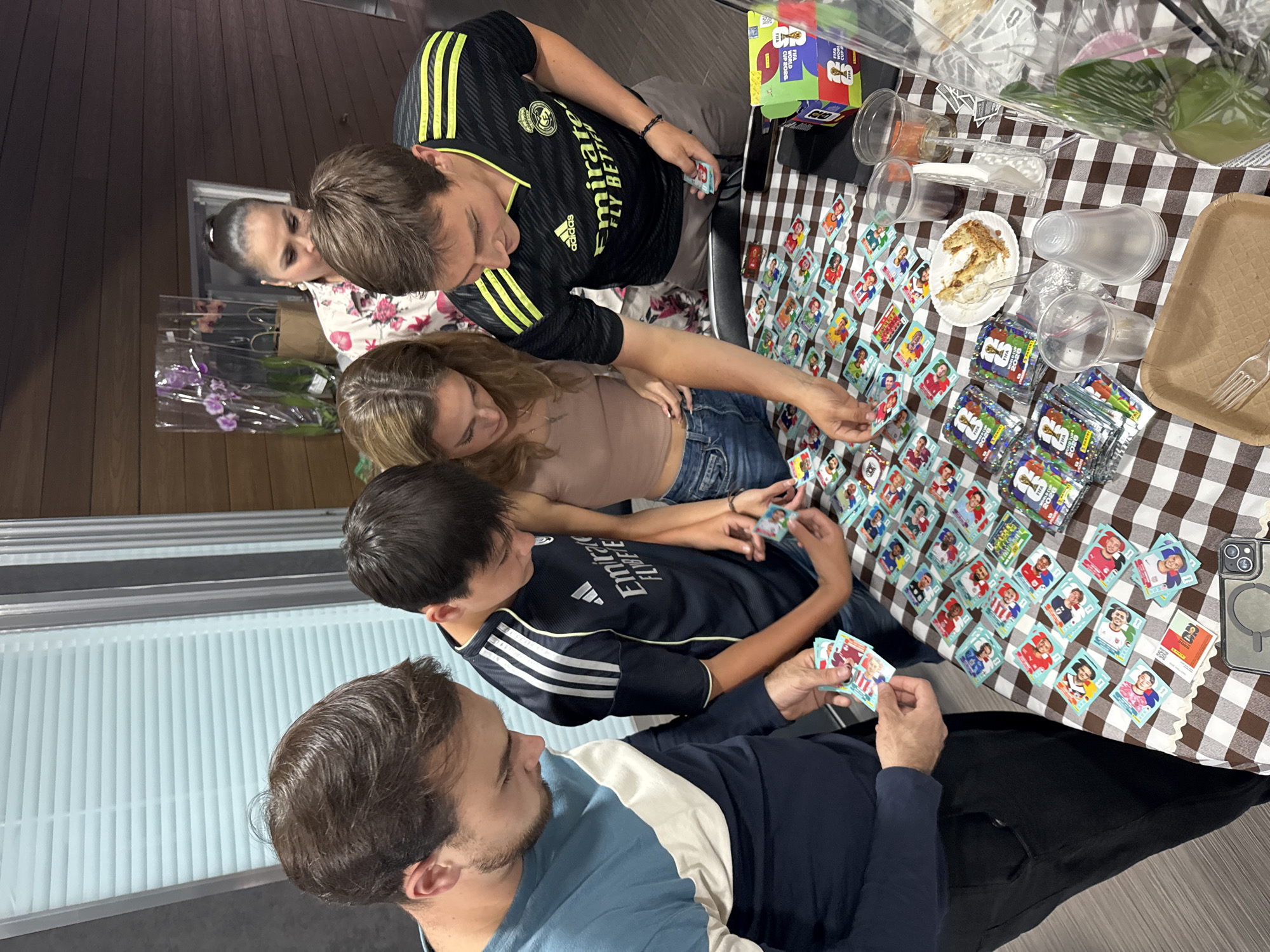 A group of five people are gathered around a table covered with a brown and white checkered tablecloth. The table is cluttered with soccer trading cards, food remnants on a plate, cups, unopened packs of trading cards, and a phone. One person is wearing a black soccer jersey with gold stripes and the text "Emirates FLY BETTER", while another wears a black soccer jersey with white stripes. Another individual is wearing a light blue shirt, while a woman wears a tan top and jeans. The location is likely indoors and based on the request, the image was taken in San Bernardino Tlaxcalancingo, Mexico. The people are focused on sorting and trading soccer cards.