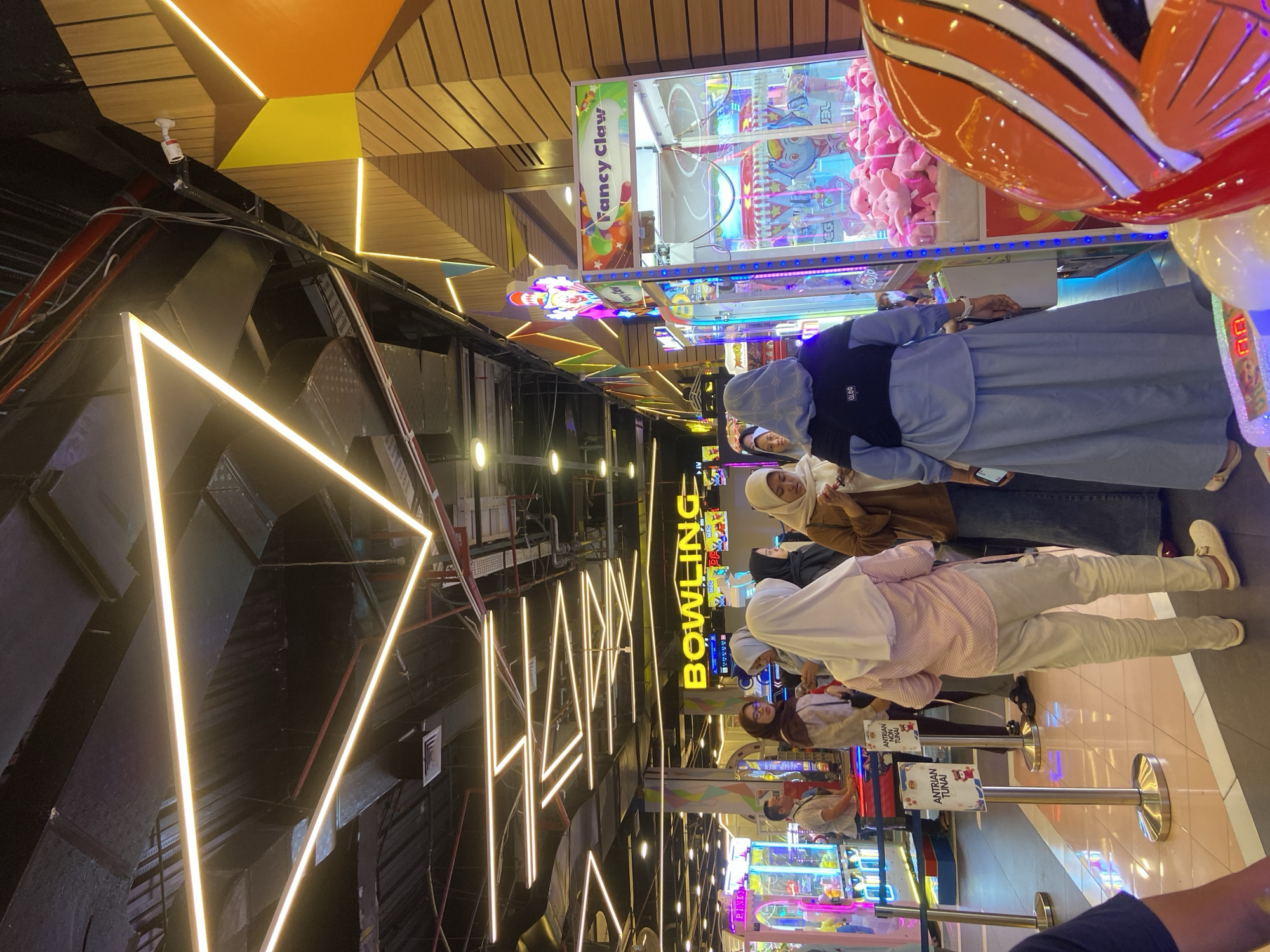The image captures a lively scene inside an entertainment center in Sidoarjo, Indonesia, likely a mall or a dedicated arcade. The environment is characterized by bright, modern lighting, with prominent triangular neon yellow light fixtures crisscrossing the ceiling. The walls feature a combination of wooden panels and colorful geometric designs.
In the foreground, a person wearing a light blue outer garment over a grey dress is seen from behind, looking towards a "Fancy Claw" arcade game filled with pink plush toys. To the left of this game, a row of other arcade machines, including what appears to be a "Pinky" game, are visible.
Further into the scene, a group of individuals, predominantly women wearing hijabs, are interacting. One woman in a light pink striped shirt and light-colored pants stands facing away from the camera, while others are engaged in conversation or looking at their phones. Two small signs with Indonesian text, possibly indicating queues or information, are positioned near the group. In the background, a large sign with the word "BOWLING" in yellow letters is clearly visible, suggesting the presence of a bowling alley. The overall atmosphere is one of active entertainment and social gathering.